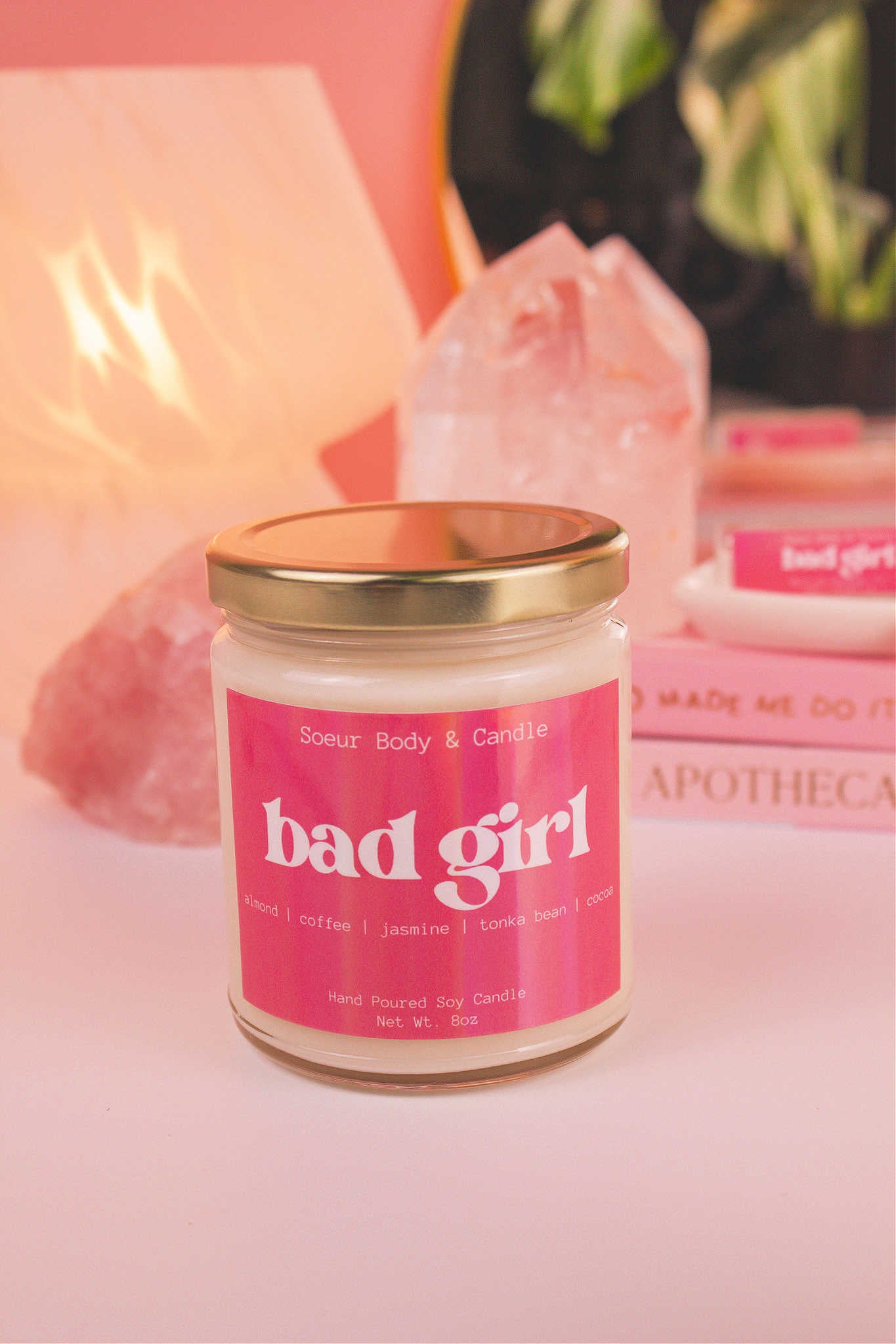 Bad Girl candle displayed on a table with books, a lamp, plant, and crystals in the background. Featuring notes of almond, coffee, jasmine, tonka bean, and cocoa.