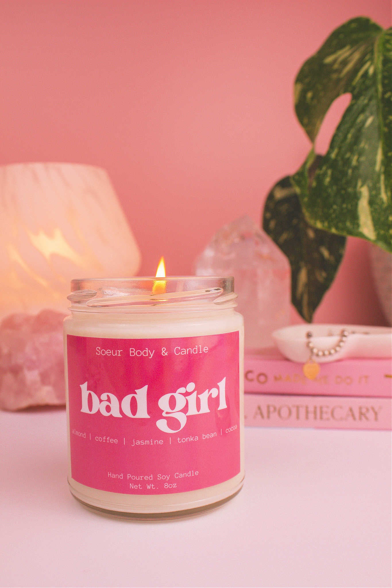 Lit Bad Girl candle with a jewelry dish on top of books, plant, lamp, and crystals in the background, showcasing its bold almond, coffee, jasmine, tonka bean, and cocoa aroma.