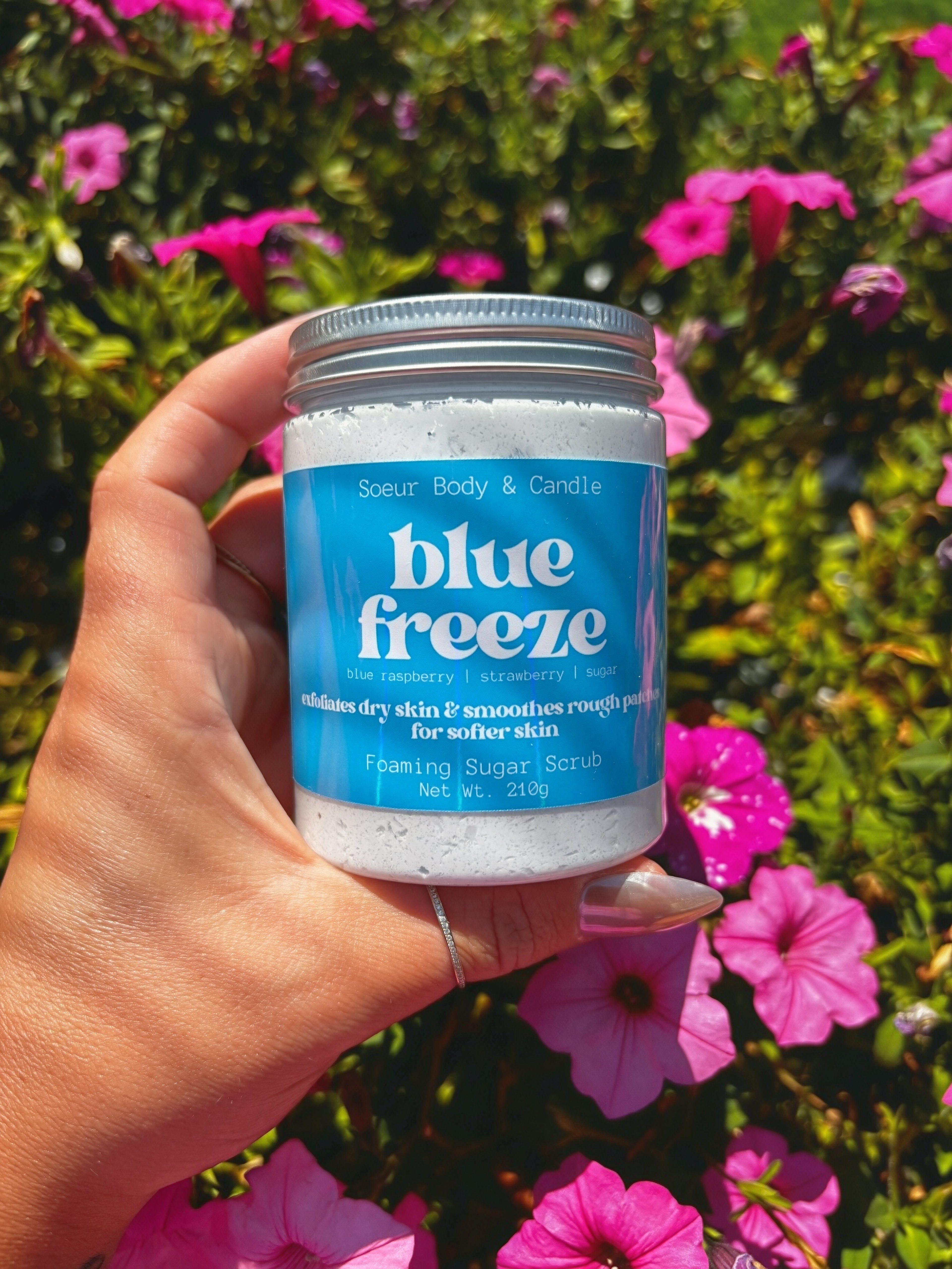 A hand holding a jar of Soeur Body and Candle Blue Freeze Foaming Sugar Scrub against a background of green foliage and pink flowers.