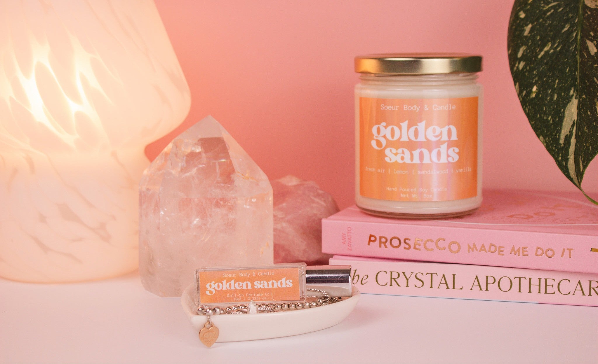 Golden Sands perfume oil displayed in a jewelry dish, with a matching candle and a stack of books in the background, set against a lamp, plant, and crystals. Fragrance notes: fresh air, lemon, sandalwood, and vanilla.