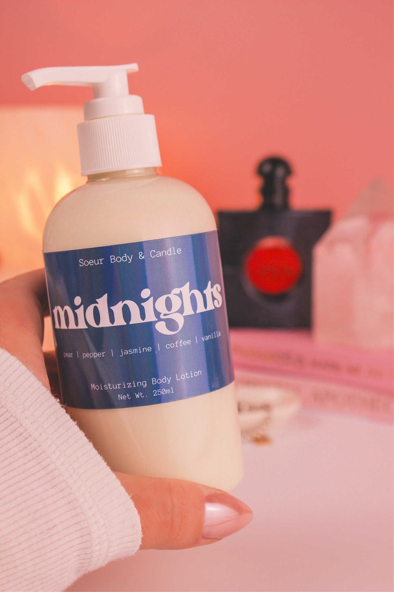 Close-up of a hand holding the Midnights body lotion, highlighting its smooth texture and exotic scent of pear, pepper, jasmine, coffee, and vanilla. There are books, a jewelry dish, lamp, perfume bottle, and crystals softly blurred in the background.