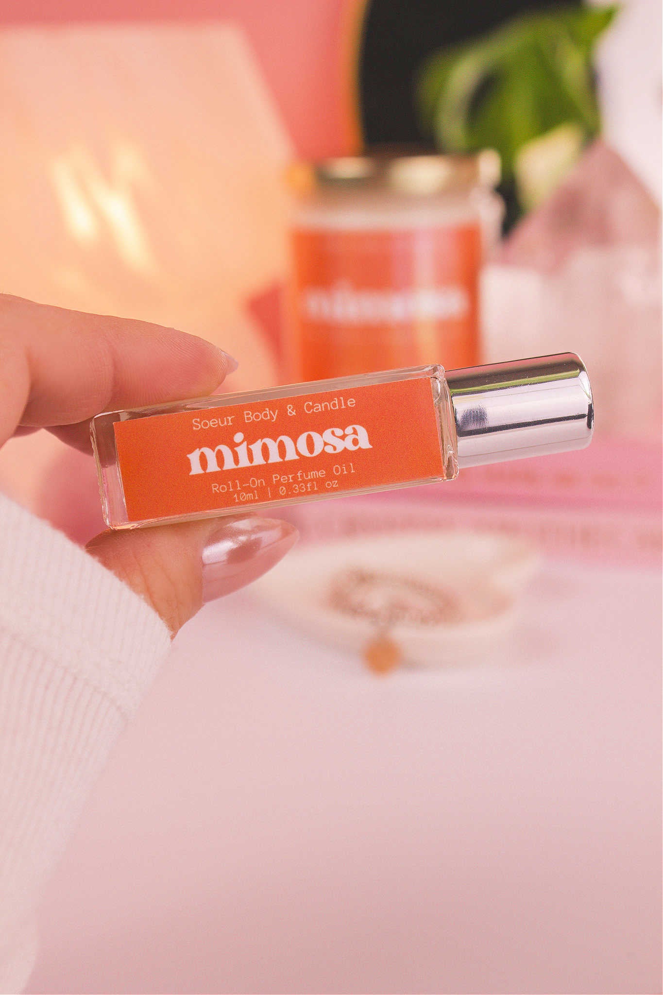 Close-up of Mimosa roll-on perfume held in hand with matching candle in the background, along with books, a lamp, and crystals, adding a cozy vibe. Featuring a blend of orange, neroli, honeysuckle, and vanilla.