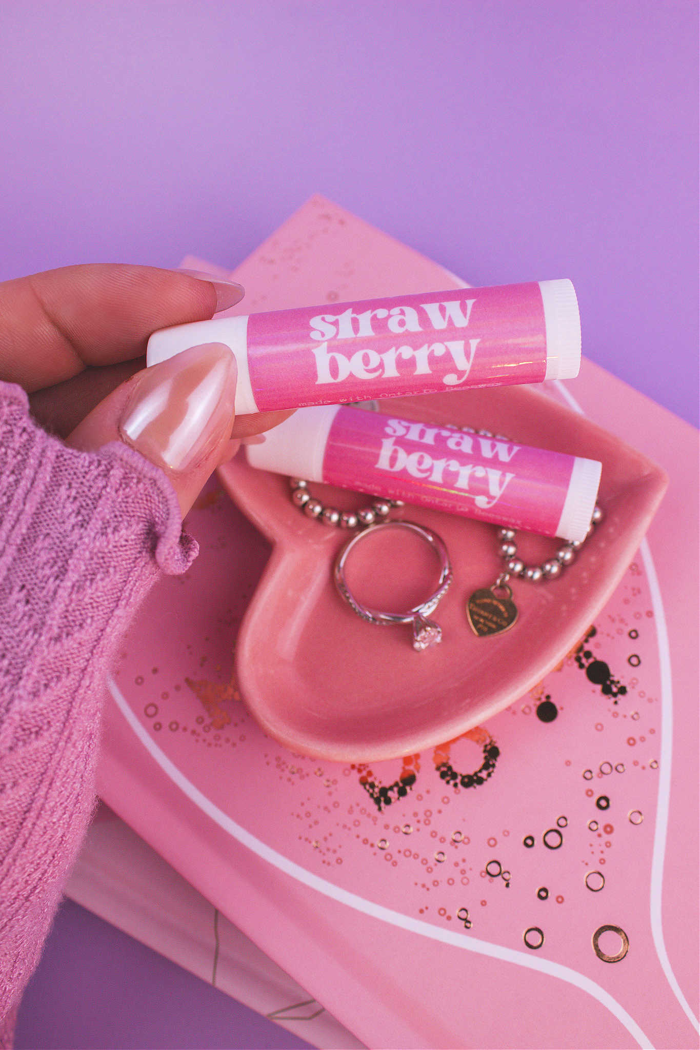 Strawberry lip balm held in hand with matching lip balm, ring, and bracelet displayed in a jewelry dish on top of books in the background.