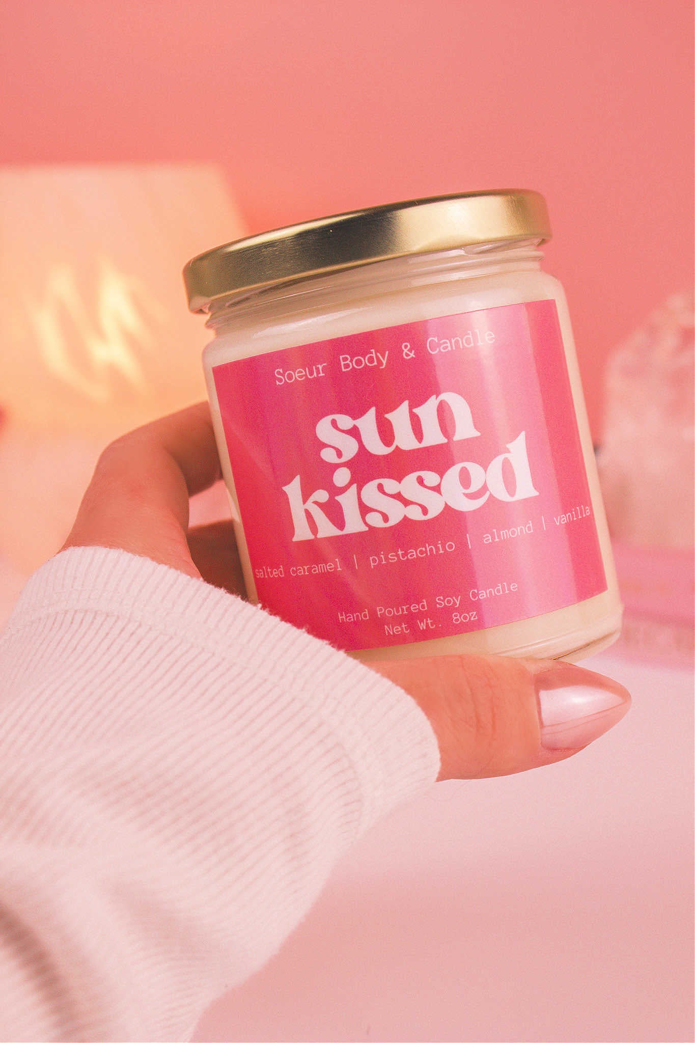 Close-up of a hand holding the Sun Kissed candle, showcasing its design and fragrance notes of salted caramel, pistachio, almond, and vanilla.