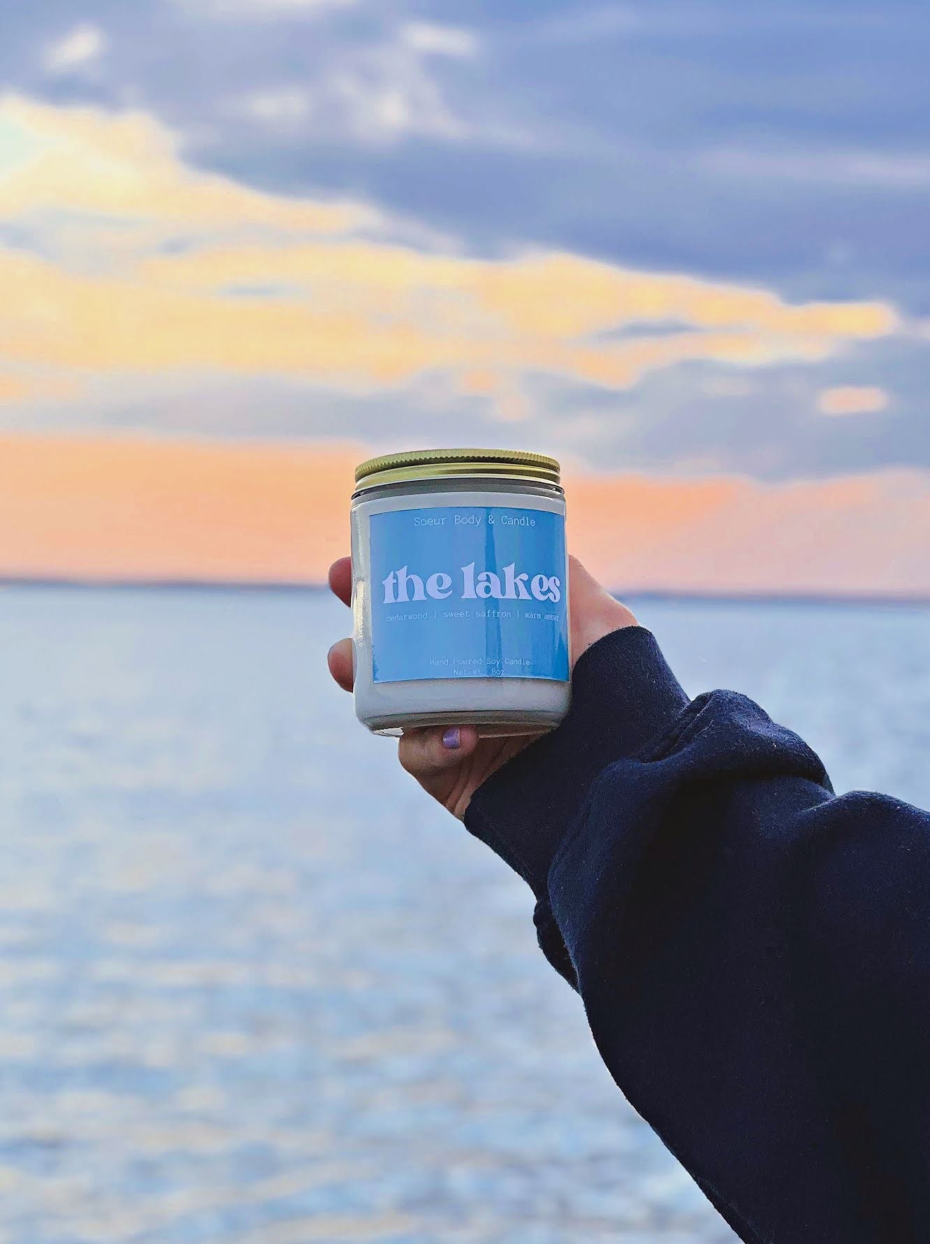 The Lakes candle held in hand, with a lake and a peach coloured sunset in the background. Fragrance notes: cedarwood, sweet saffron, and warm amber.