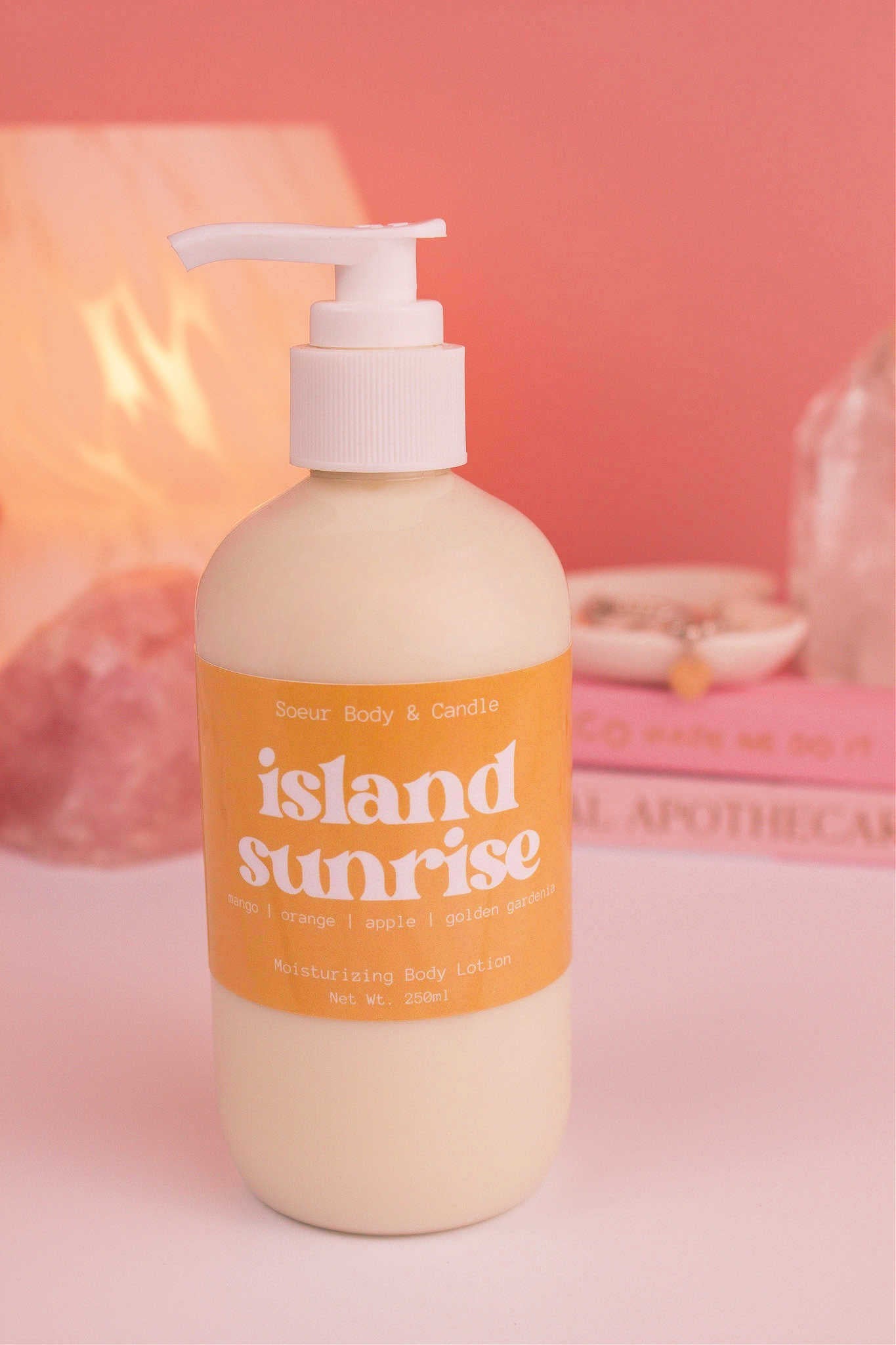 Island Sunrise body lotion with a stack of books, a jewelry dish, lamp, and crystals in the background; fragrance notes: mango, orange, apple, and golden gardenia.