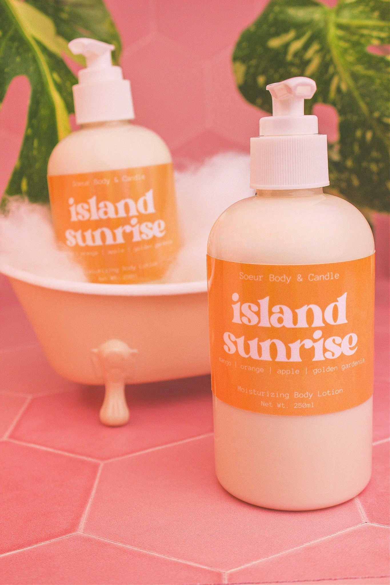 Display of two Island Sunrise body lotions in a small tub with bubbles and plants in the background, evoking a relaxing vibe with mango, orange, apple, and golden gardenia notes.