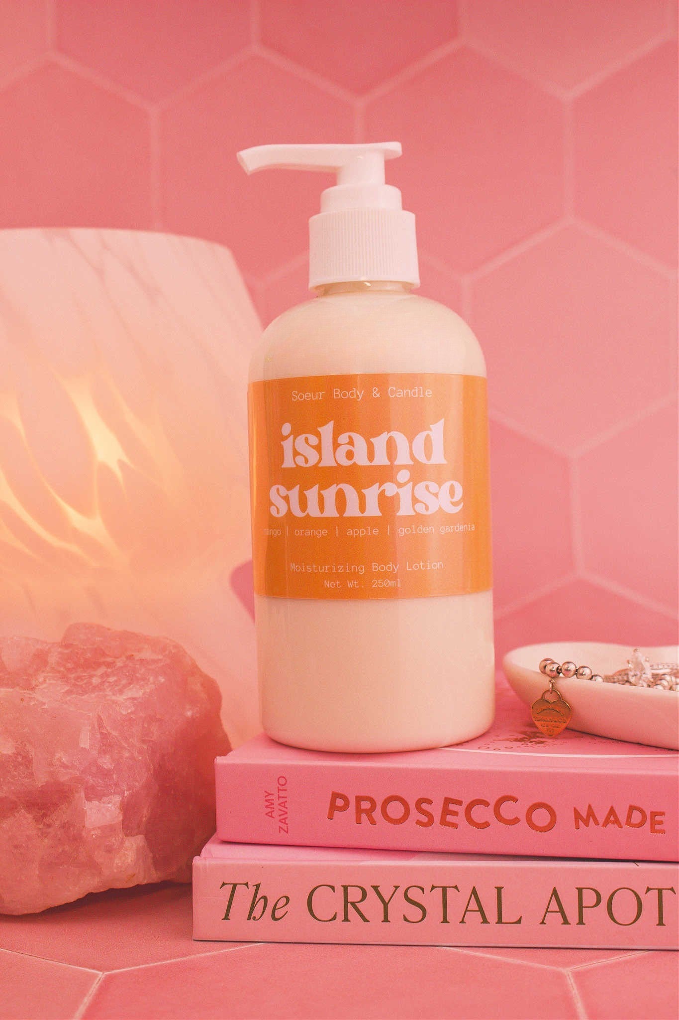 Close-up of the Island Sunrise body lotion on a stack of books with a jewelry dish, lamp, and crystal in the background; fragrance notes: mango, orange, apple, and golden gardenia.