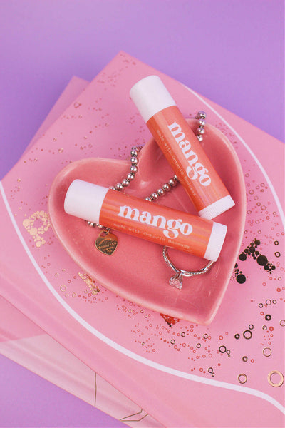 Two Mango flavoured lip balms displayed in a jewelry dish with a ring and bracelet, sitting on top of books.