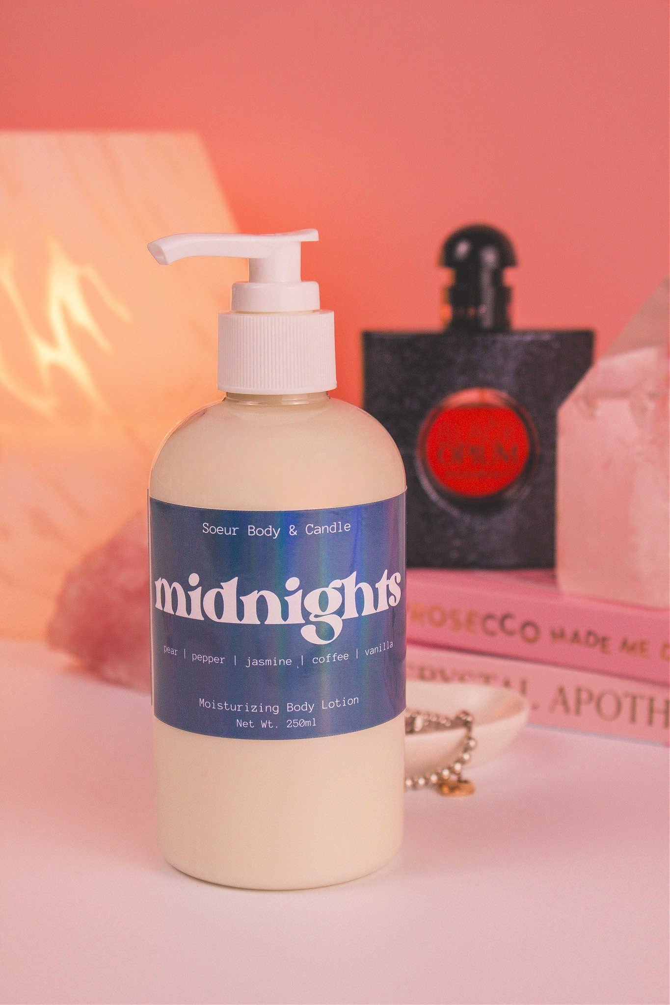 Midnights body lotion with a stack of books, a jewelry dish, lamp, perfume bottle, and crystals in the background; fragrance notes: pear, pepper, jasmine, coffee, and vanilla.