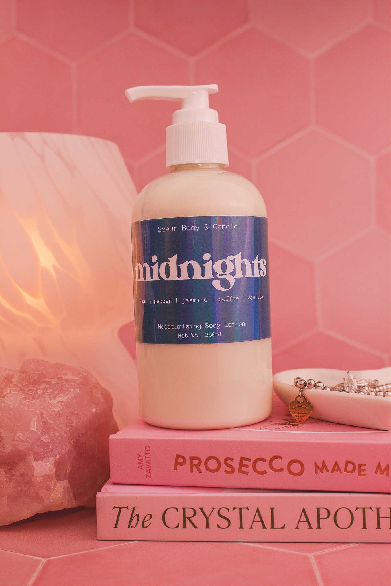 Close-up of the Midnights body lotion on a stack of books with a jewelry dish, lamp, and crystals in the background; fragrance notes: pear, pepper, jasmine, coffee, and vanilla.