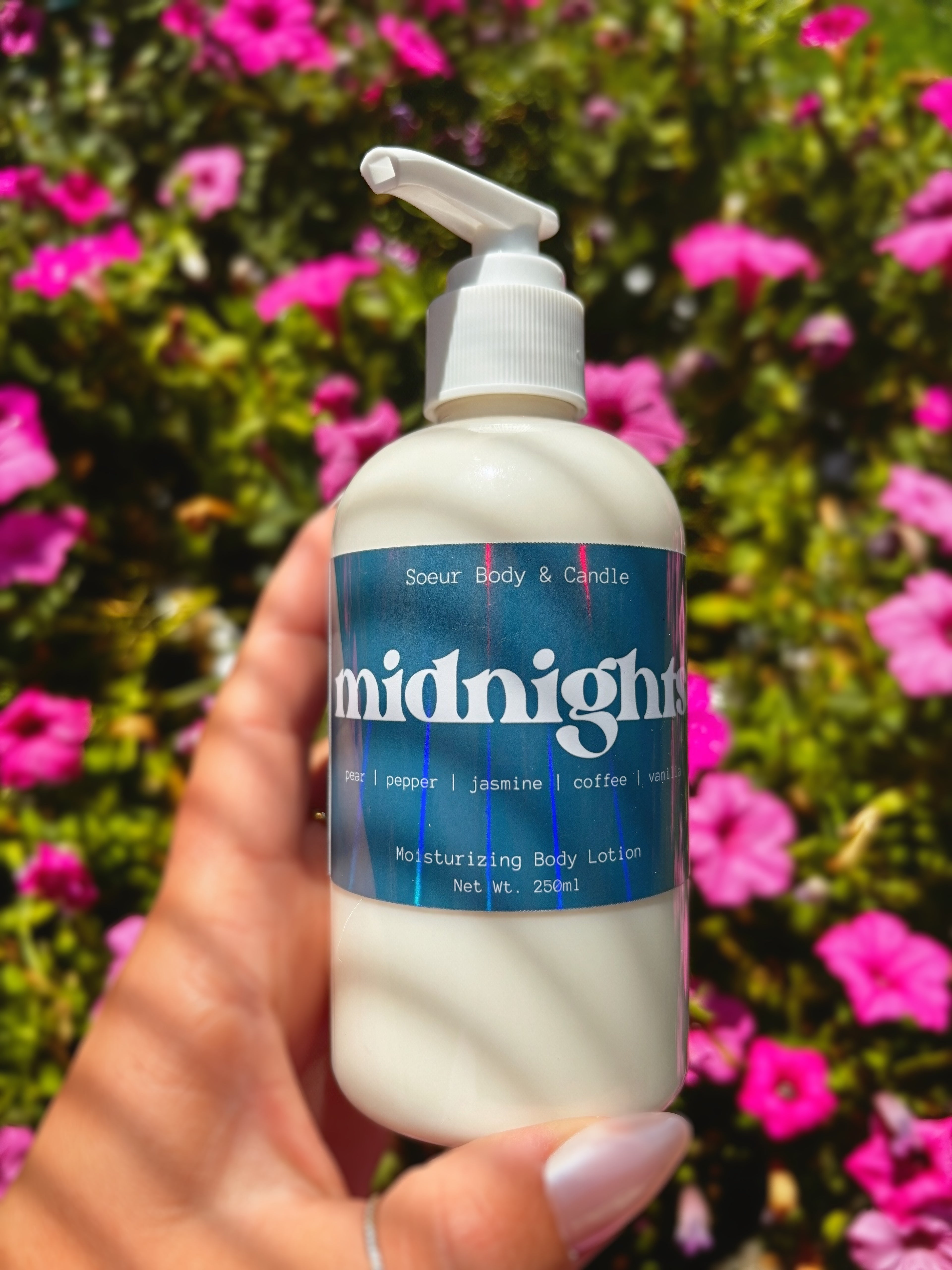 A hand holding a bottle of Midnights Body Lotion against a background of green foliage and pink flowers. The bottle is white with a blue label and a white pump dispenser.