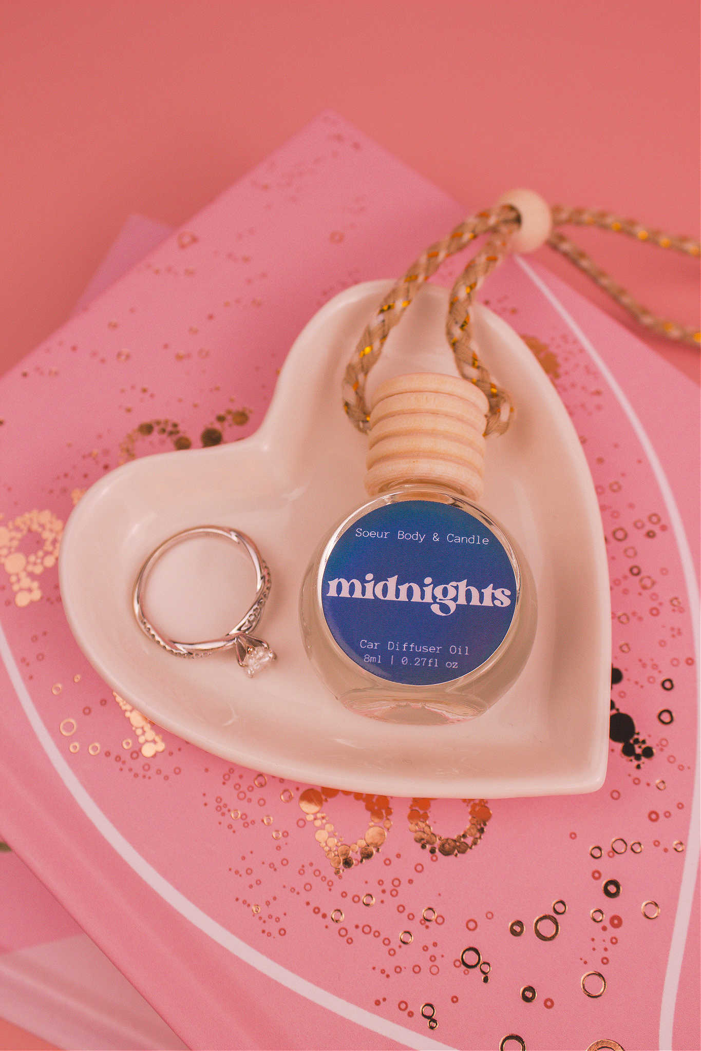 Midnights car diffuser in a jewelry dish with a ring beside it, sitting on top of books. Featuring an exotic blend of pear, pepper, jasmine, coffee, and vanilla notes.