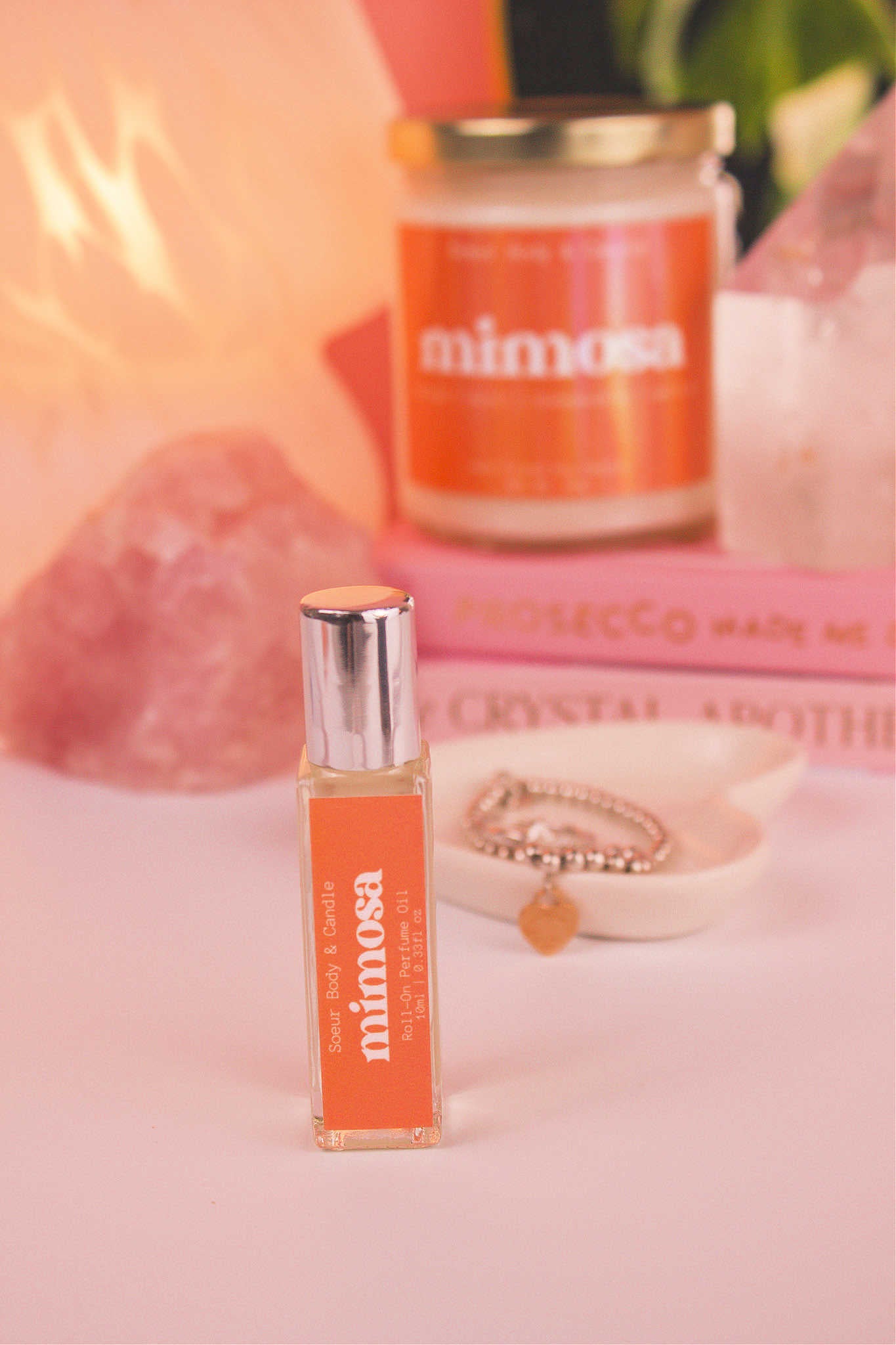 Mimosa roll-on perfume in the foreground, with a matching candle, jewelry dish, books, a lamp, and crystals in the background, adding a cozy vibe. Featuring notes of orange, neroli, honeysuckle, and vanilla.