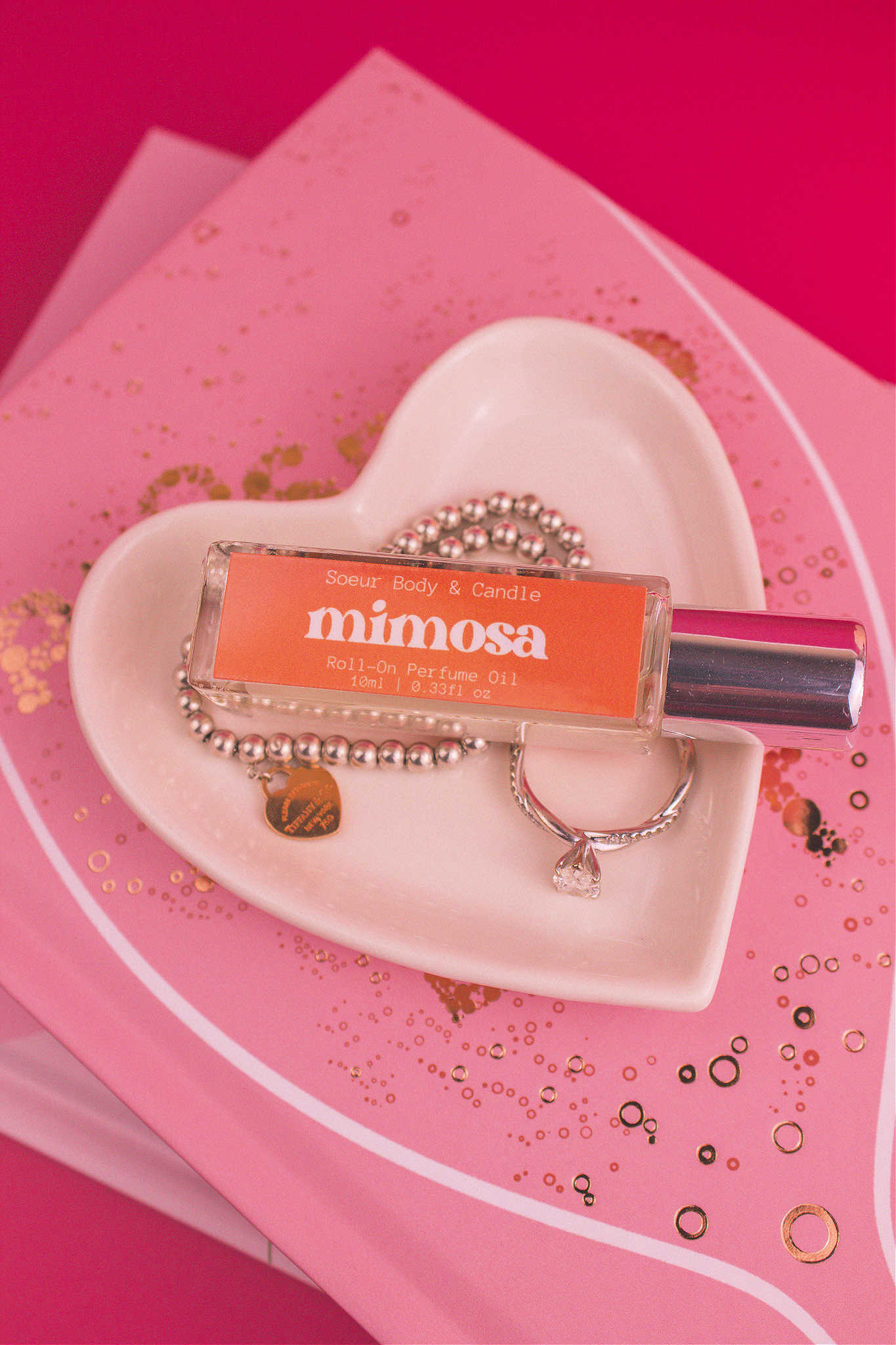 Mimosa roll-on perfume bottle in a jewelry dish beside a ring and bracelet, highlighting its fruity notes of orange, neroli, honeysuckle, and vanilla.