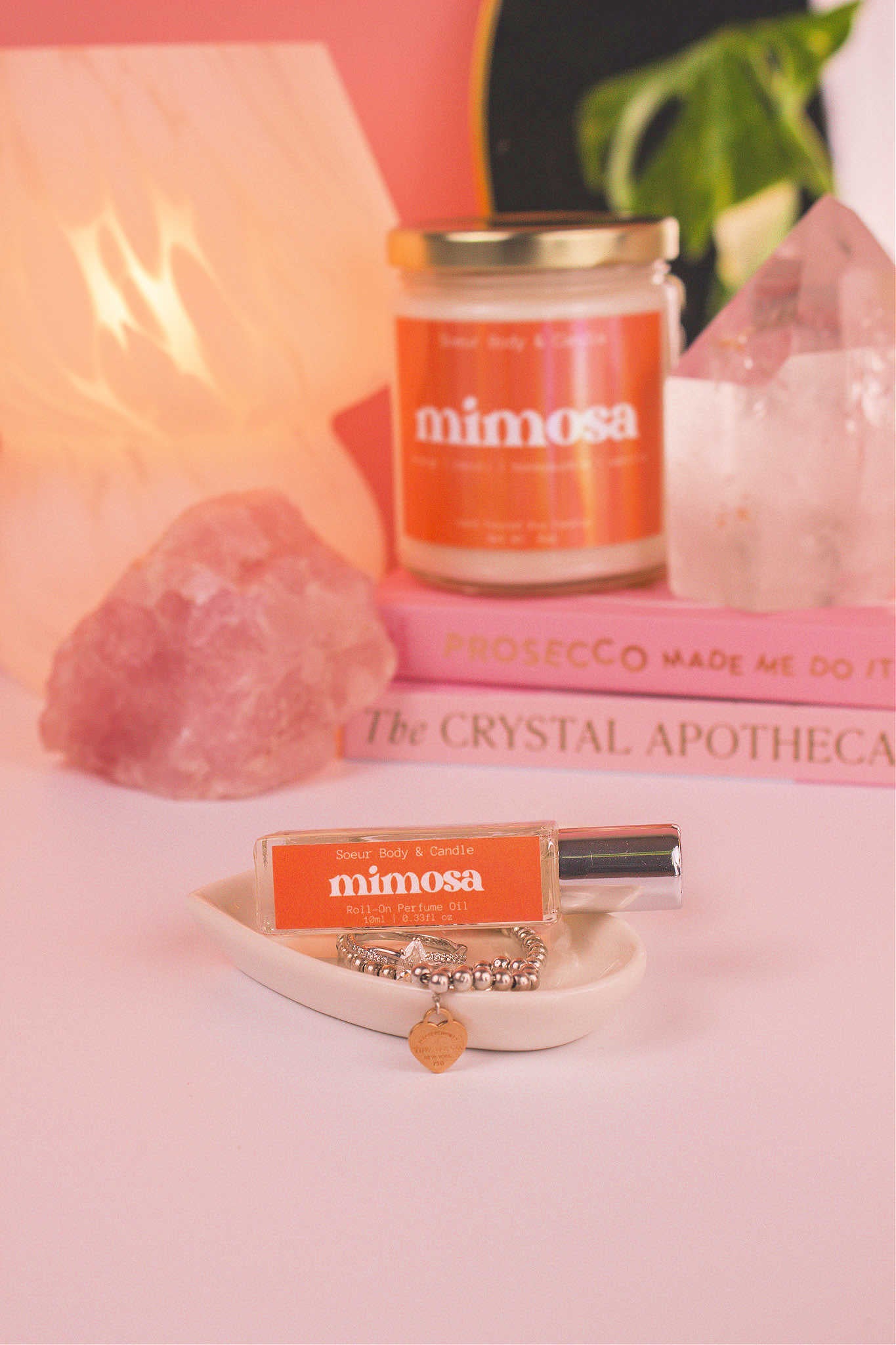 Mimosa roll-on perfume elegantly displayed in a jewelry dish with coordinating jewelry. Matching candle, books, a lamp, and crystals in the background add a cozy vibe. Featuring notes of orange, neroli, honeysuckle, and vanilla.