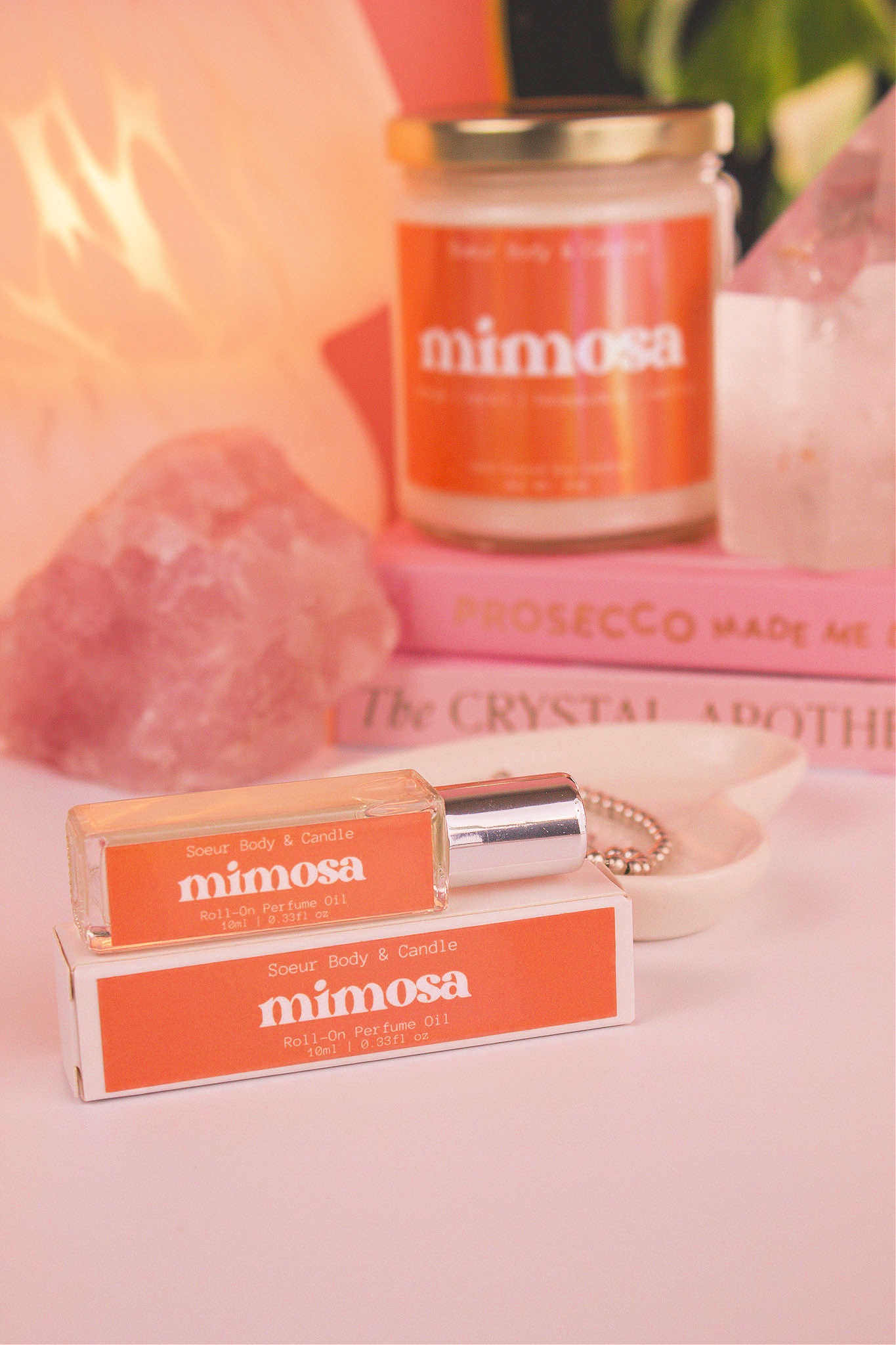 Focused shot of Mimosa roll-on perfume in the foreground with matching candle softly blurred in the background, along with books, a lamp, and crystals, adding a cozy vibe. Featuring notes of orange, neroli, honeysuckle, and vanilla.