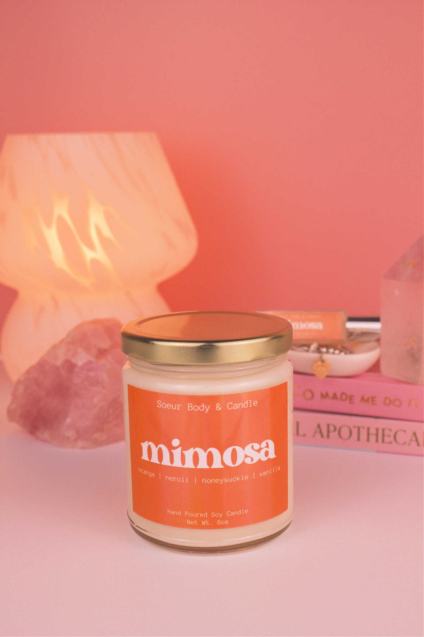 Mimosa candle with the lid on, with a stack of books and a jewelry dish in the background, set against a lamp and crystals; fragrance notes: orange, neroli, honeysuckle, and vanilla.