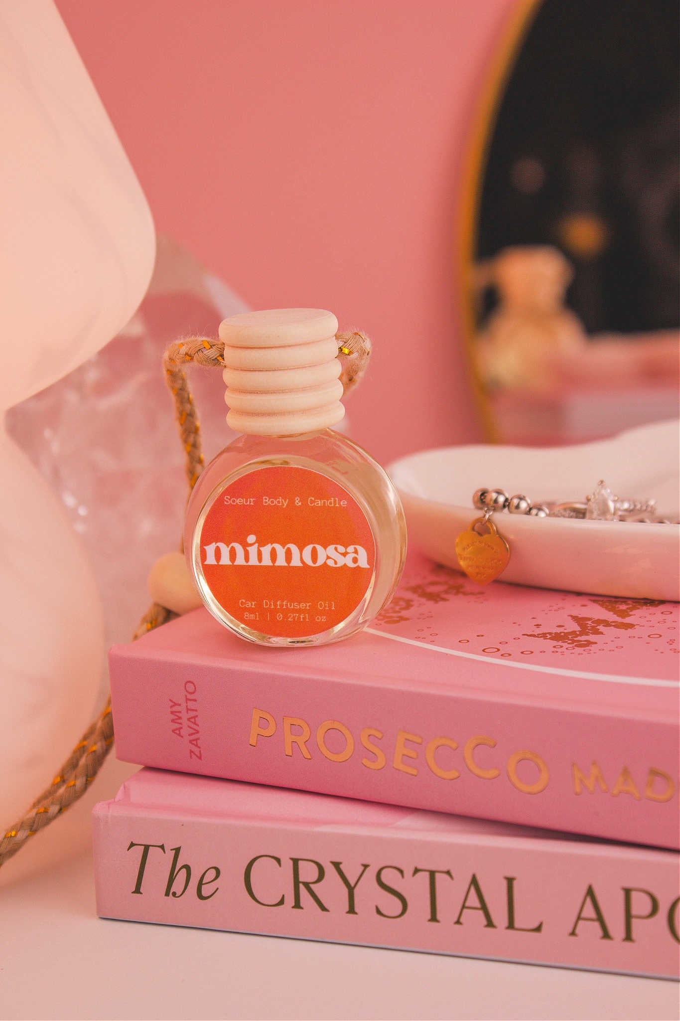Mimosa diffuser sitting on top of a stack of books with a jewelry dish in the background, set against a lamp, mirror, and crystals; fragrance notes: orange, neroli, honeysuckle, and vanilla.