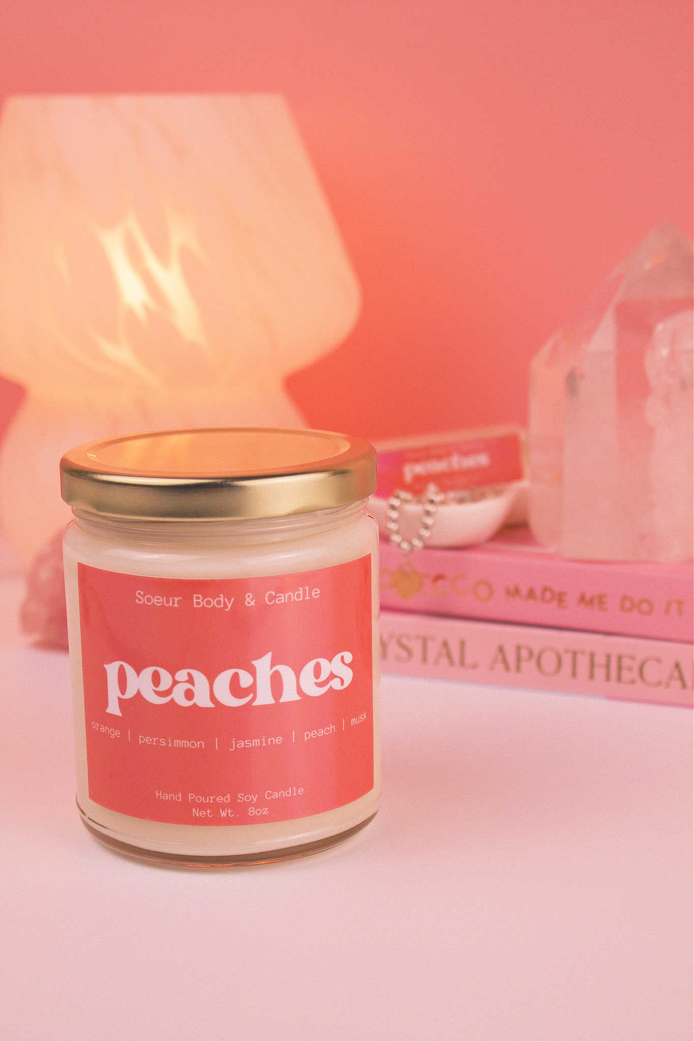 Close-up of Peaches candle with the lid on, with a stack of books and a jewelry dish in the background, set against a lamp and crystals; fragrance notes: orange, persimmon, jasmine, peach, and musk.