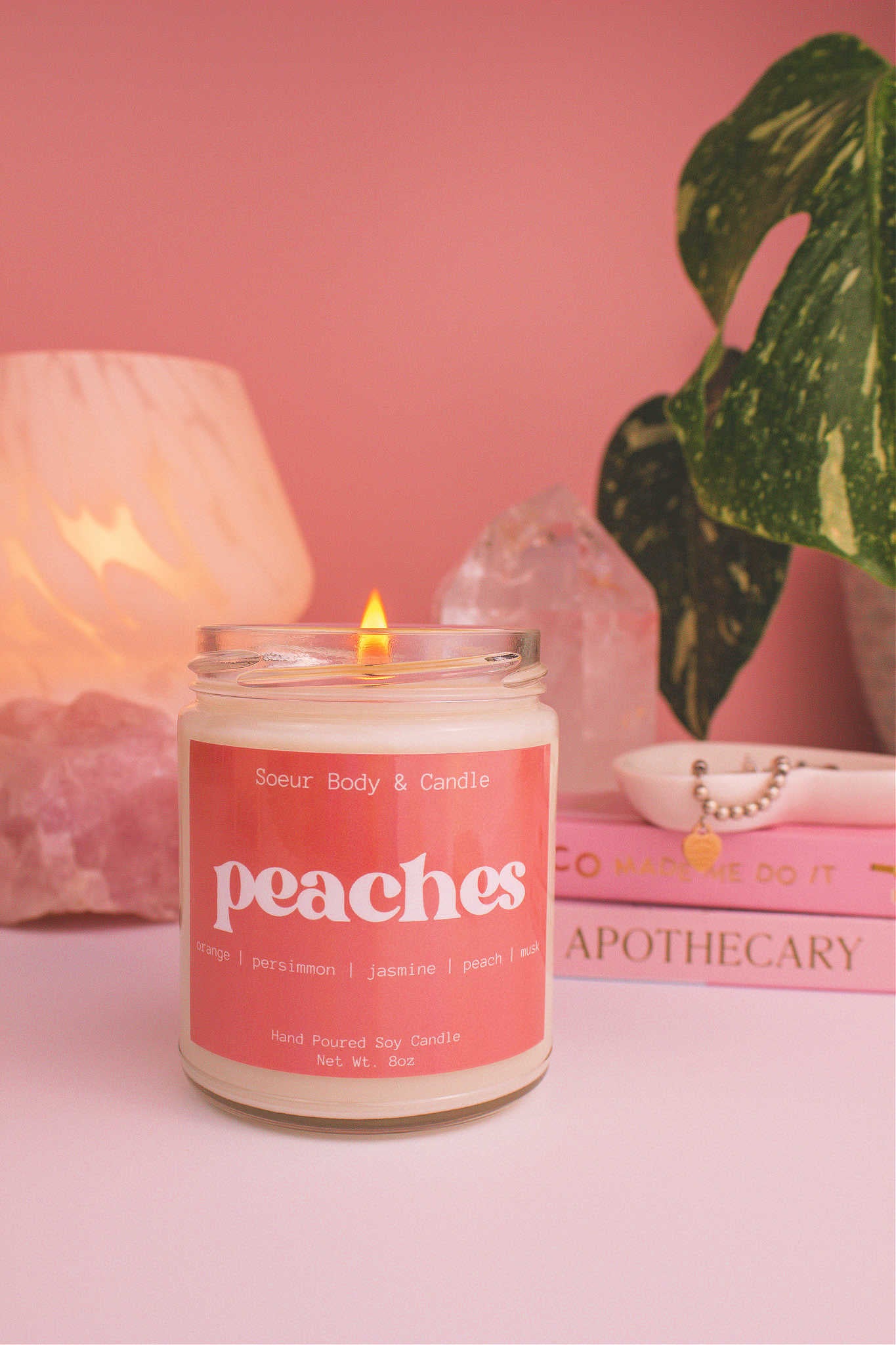 Peaches candle lit, with a stack of books and a jewelry dish in the background, set against a lamp, plant, and crystals; fragrance notes: orange, persimmon, jasmine, peach, and musk.