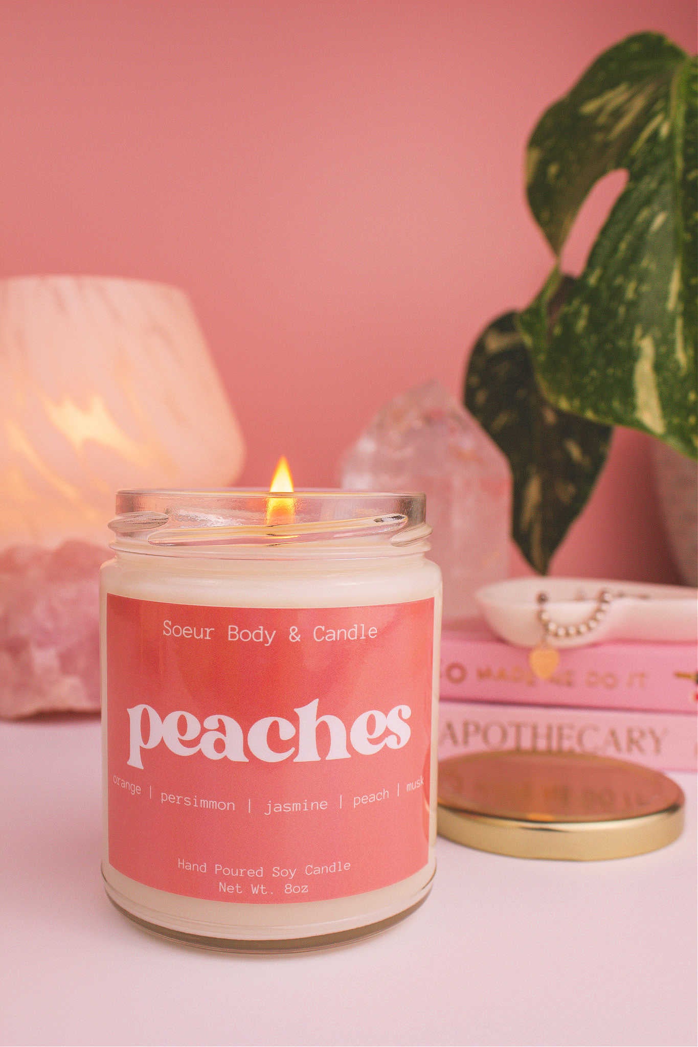 Close-up of Peaches candle lit, with a stack of books and a jewelry dish in the background, set against a lamp, plant, and crystals; fragrance notes: orange, persimmon, jasmine, peach, and musk.