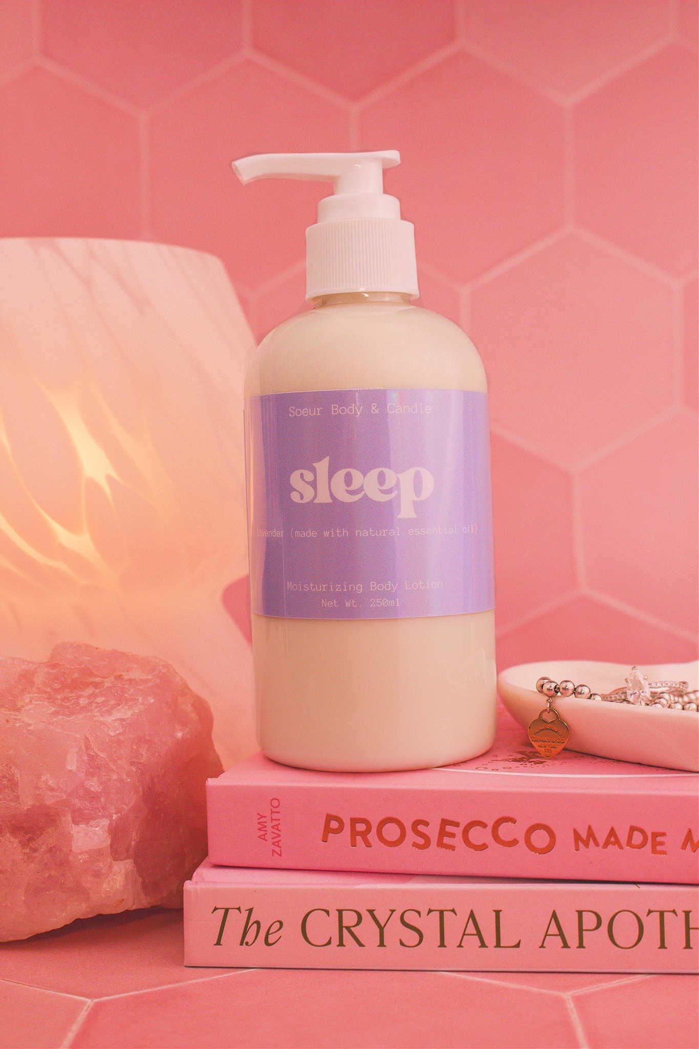 Close-up of the Sleep body lotion on a stack of books with a jewelry dish, lamp, and crystal in the background; scented with lavender essential oil.