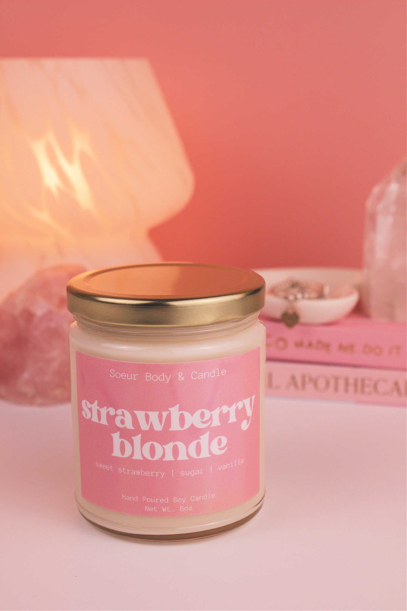 Strawberry Blonde candle with the lid on, with a stack of books and a jewelry dish in the background, set against a lamp and crystals; fragrance notes: sweet strawberry, sugar, and vanilla.