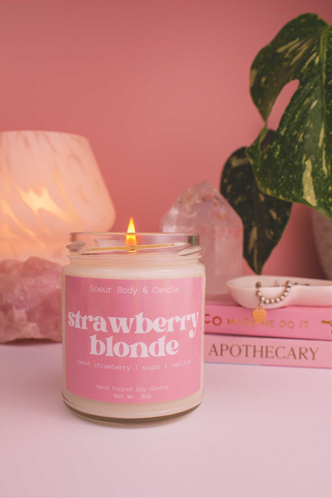 Strawberry Blonde candle lit, with a stack of books and a jewelry dish in the background, set against a lamp, plant, and crystals; fragrance notes: sweet strawberry, sugar, and vanilla.