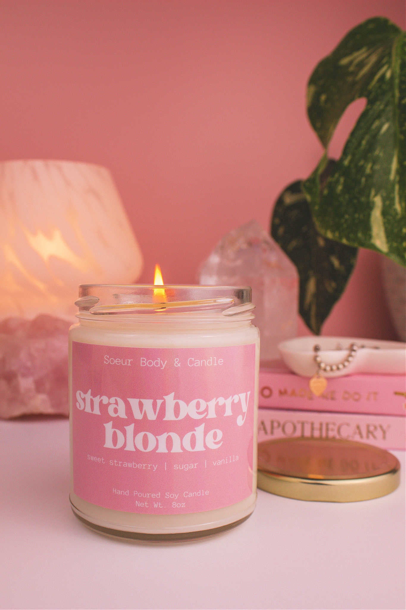Close-up of Strawberry Blonde candle lit, with a stack of books and a jewelry dish in the background, set against a lamp, plant, and crystals; fragrance notes: sweet strawberry, sugar, and vanilla.