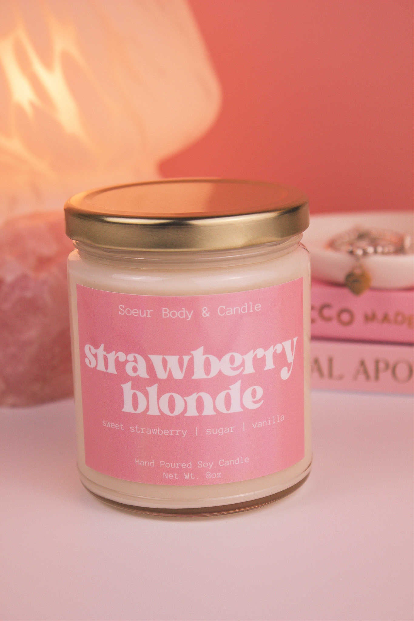 Close-up of Strawberry Blonde candle with the lid on, with a stack of books and a jewelry dish in the background, set against a lamp and crystals; fragrance notes: sweet strawberry, sugar, and vanilla.