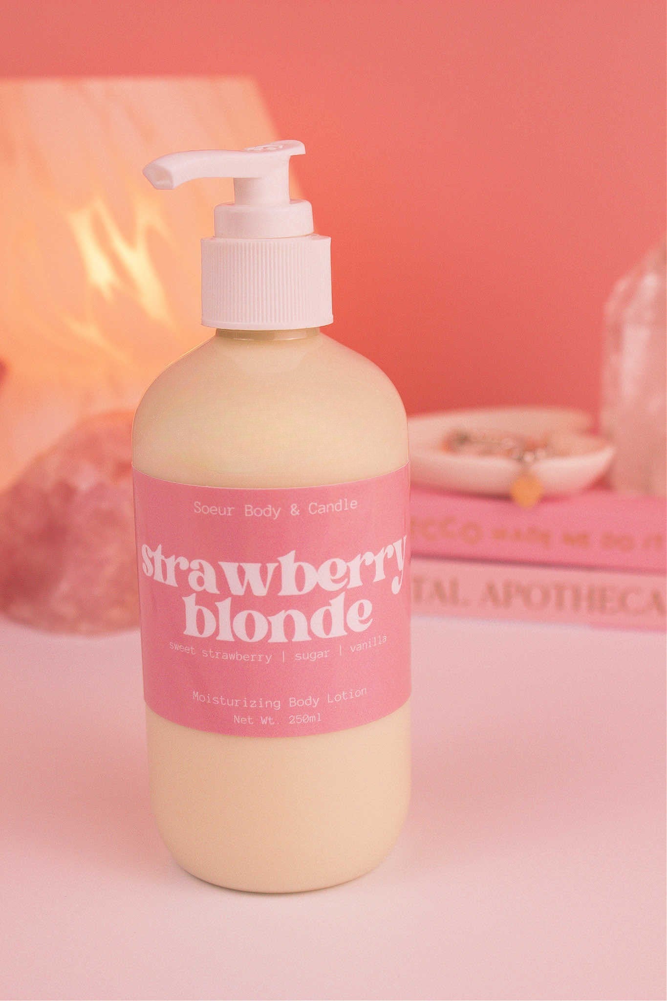 Strawberry Blonde body lotion with a stack of books, a jewelry dish, lamp, and crystals in the background; fragrance notes: sweet strawberry, sugar, and vanilla.