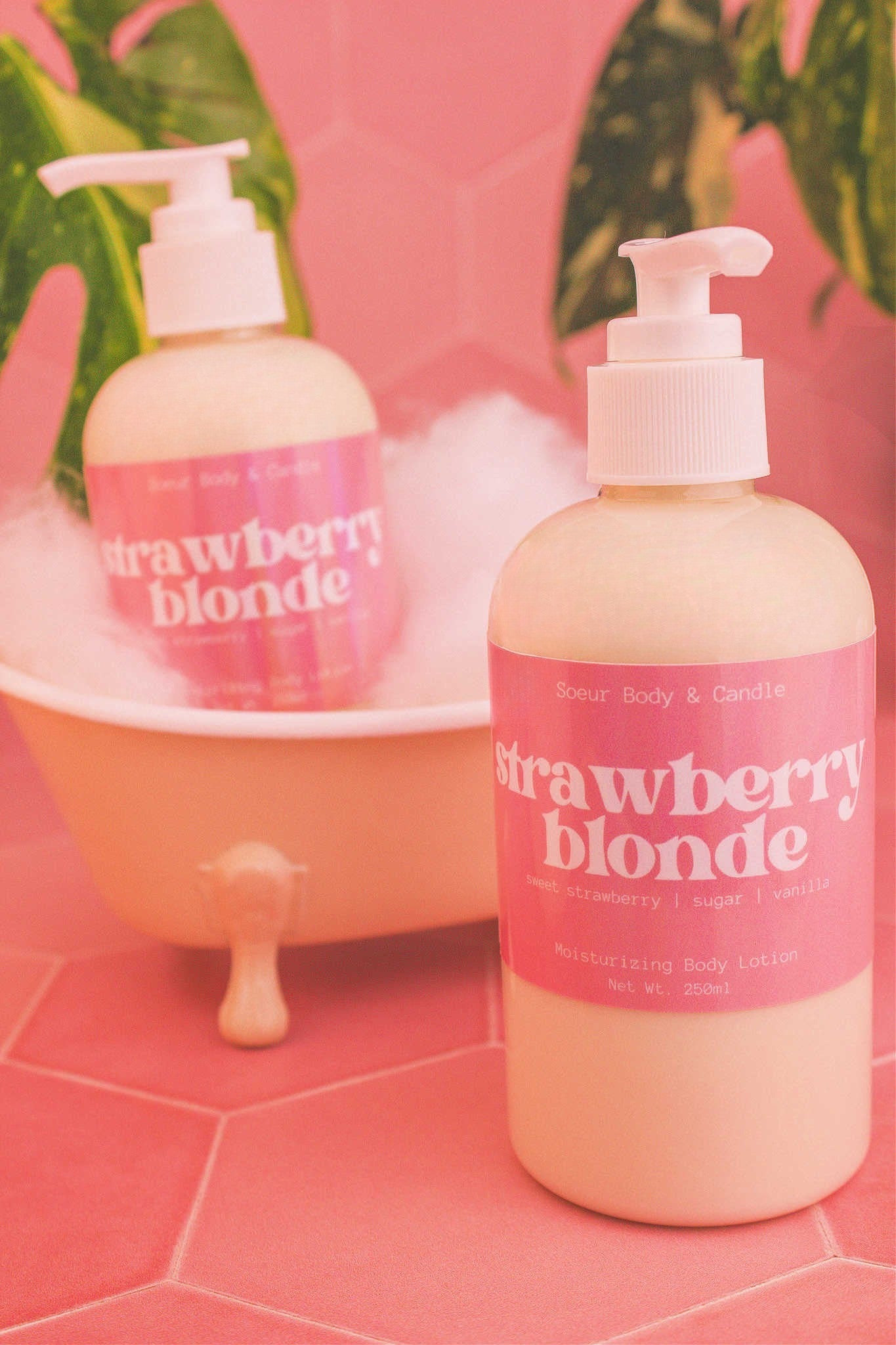 Display of two Strawberry Blonde body lotions in a small tub with bubbles and plants in the background, evoking a relaxing vibe with sweet strawberry, sugar, and vanilla notes.