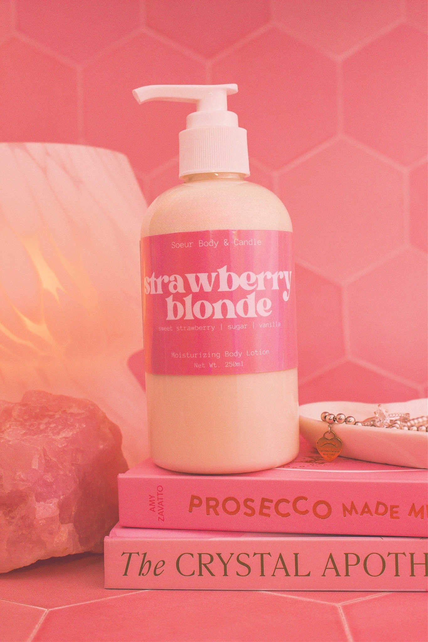 Close-up of the Strawberry Blonde body lotion on a stack of books with a jewelry dish, lamp, and crystal in the background; fragrance notes: sweet strawberry, sugar, and vanilla.