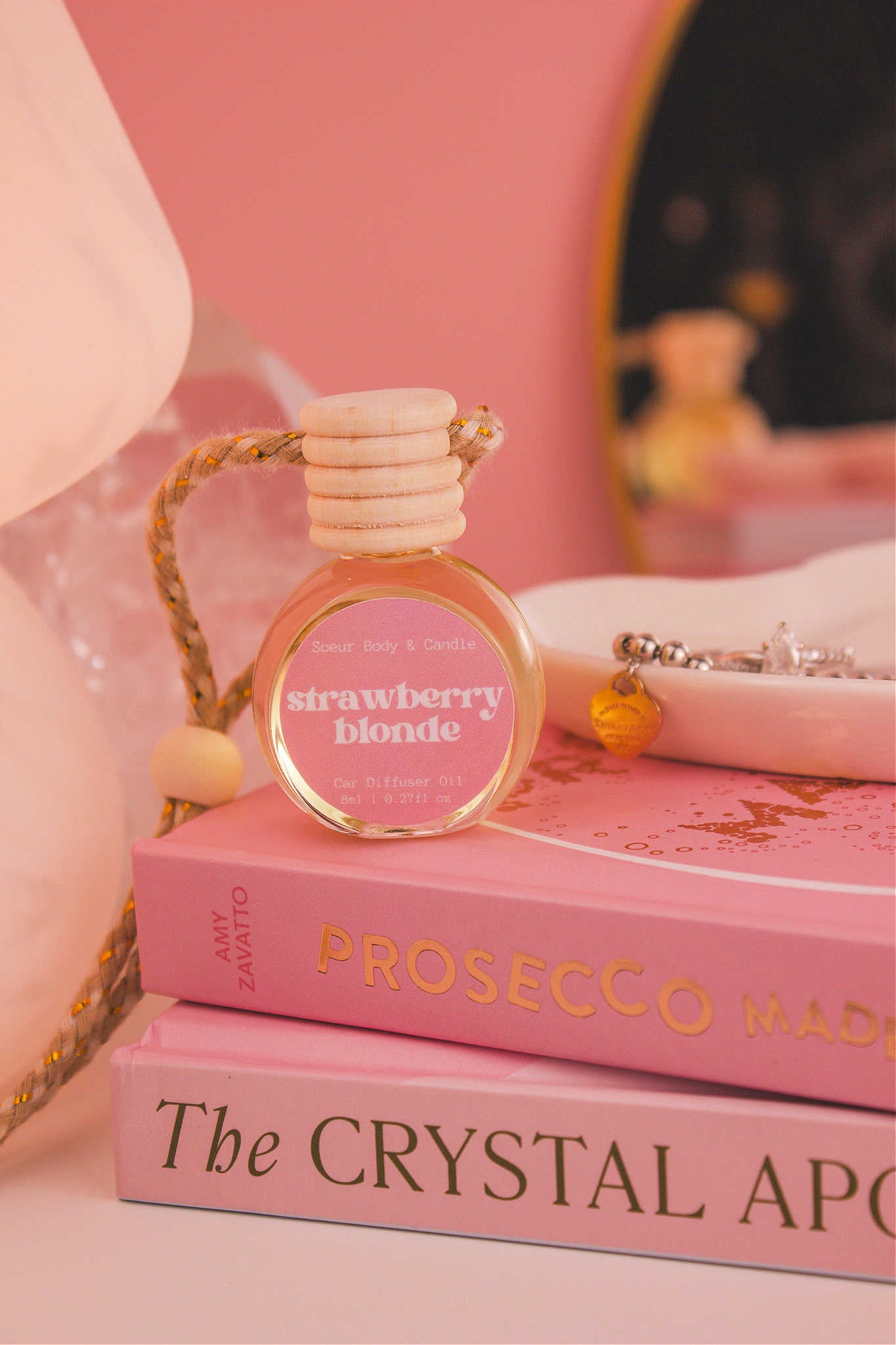 Strawberry Blonde diffuser sitting on top of a stack of books with a jewelry dish in the background, set against a lamp, mirror, and crystals; fragrance notes: sweet strawberry, sugar, and vanilla.