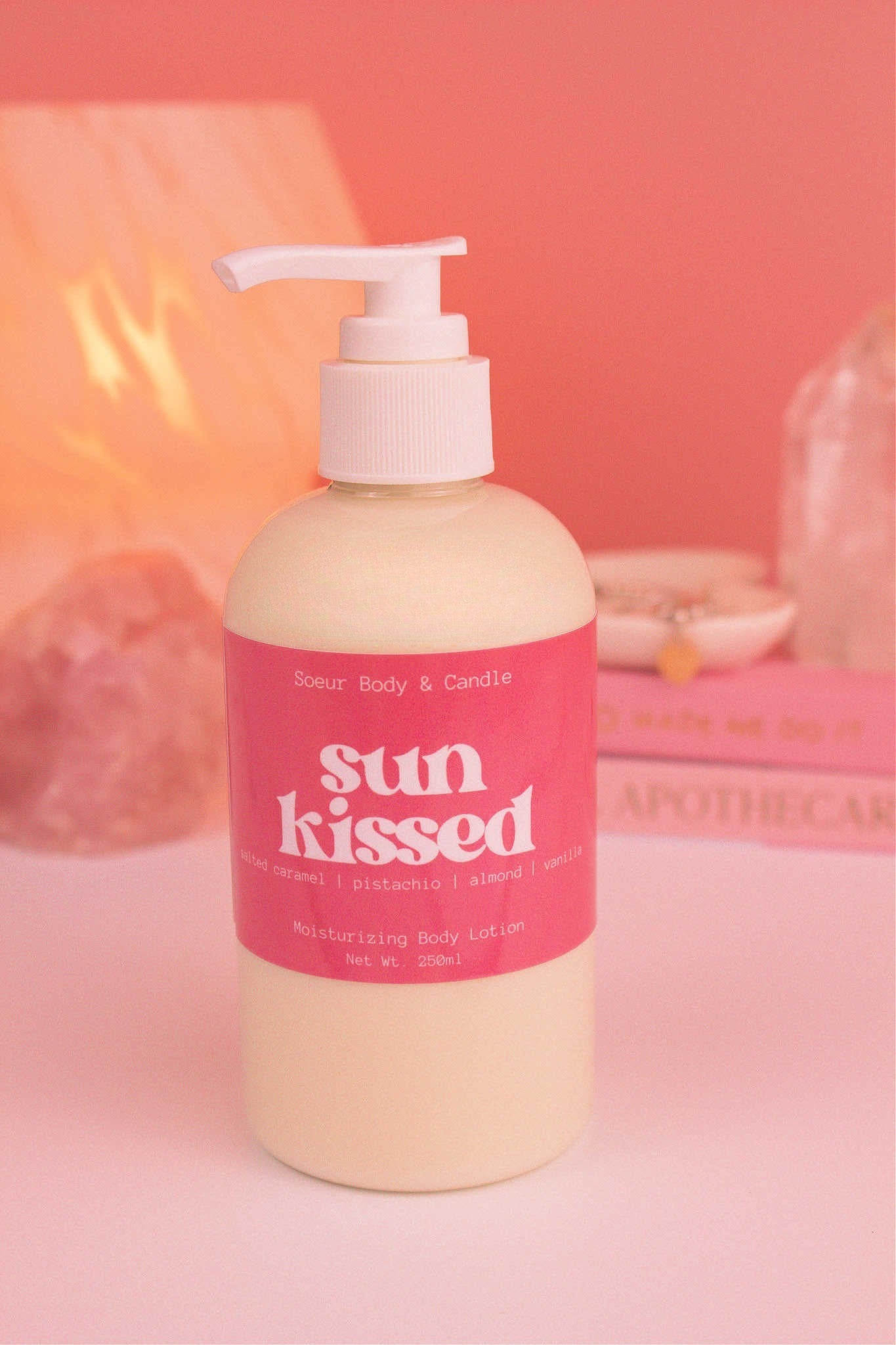 Close-up of Sun Kissed body lotion with a stack of books, a jewelry dish, lamp, and crystals in the background; fragrance notes: salted caramel, pistachio, almond, and vanilla.