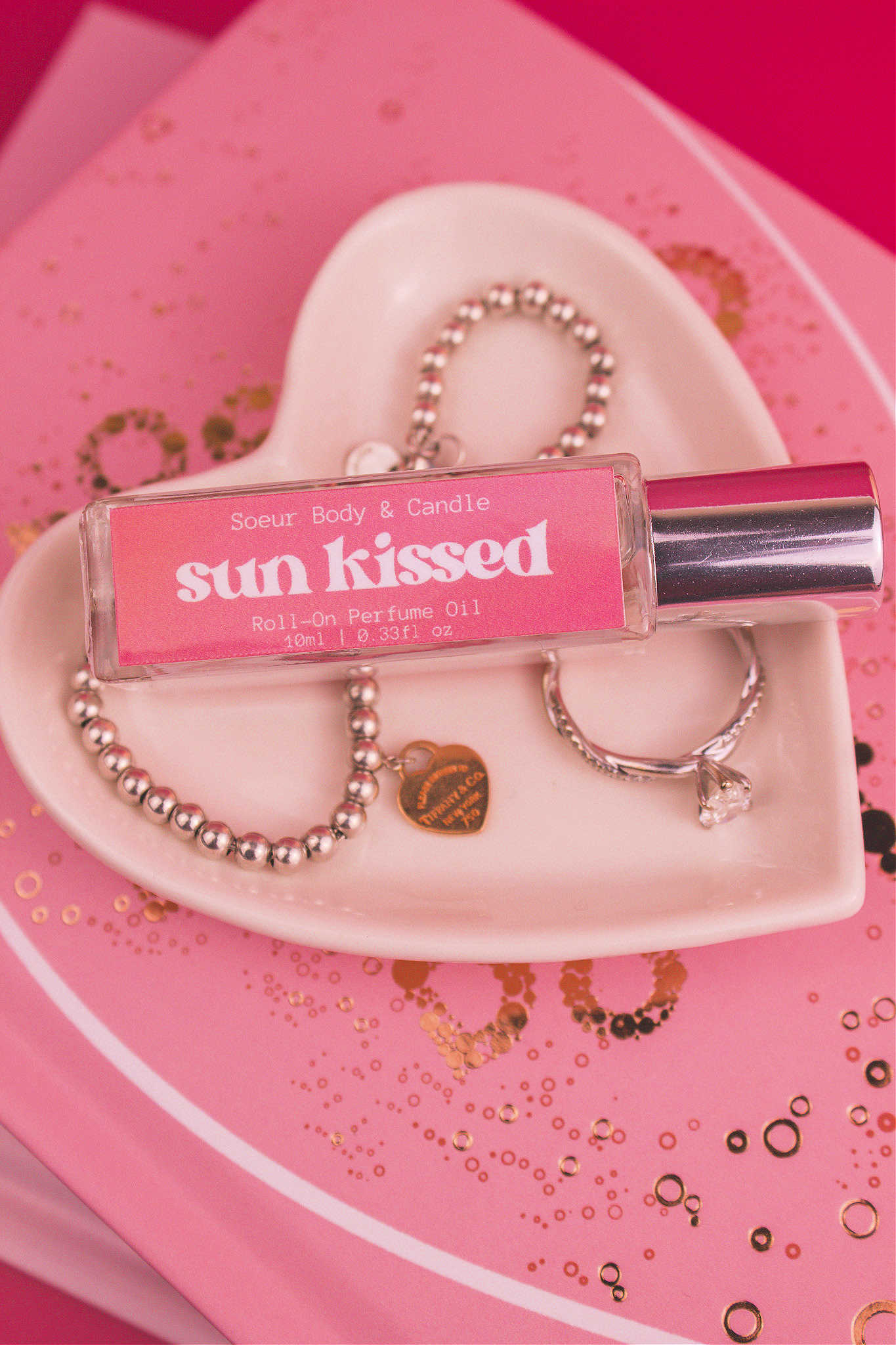 Close-up of Sun Kissed roll-on perfume bottle displayed in a jewelry dish beside a ring and bracelet, on top of books. Featuring notes of salted caramel, pistachio, almond, and vanilla.