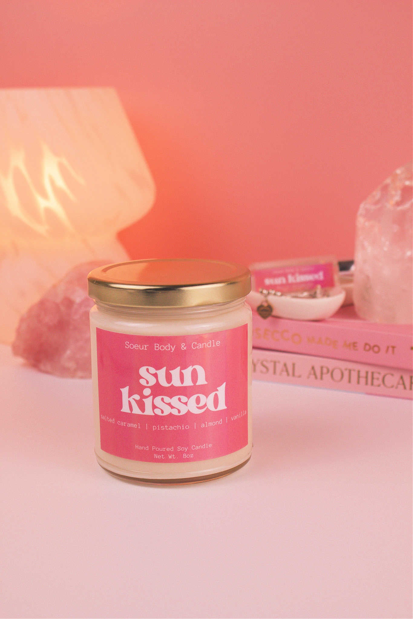 Sun Kissed candle with the lid on, with a stack of books and a jewelry dish in the background, set against a lamp and crystals; fragrance notes: salted caramel, pistachio, almond, and vanilla.