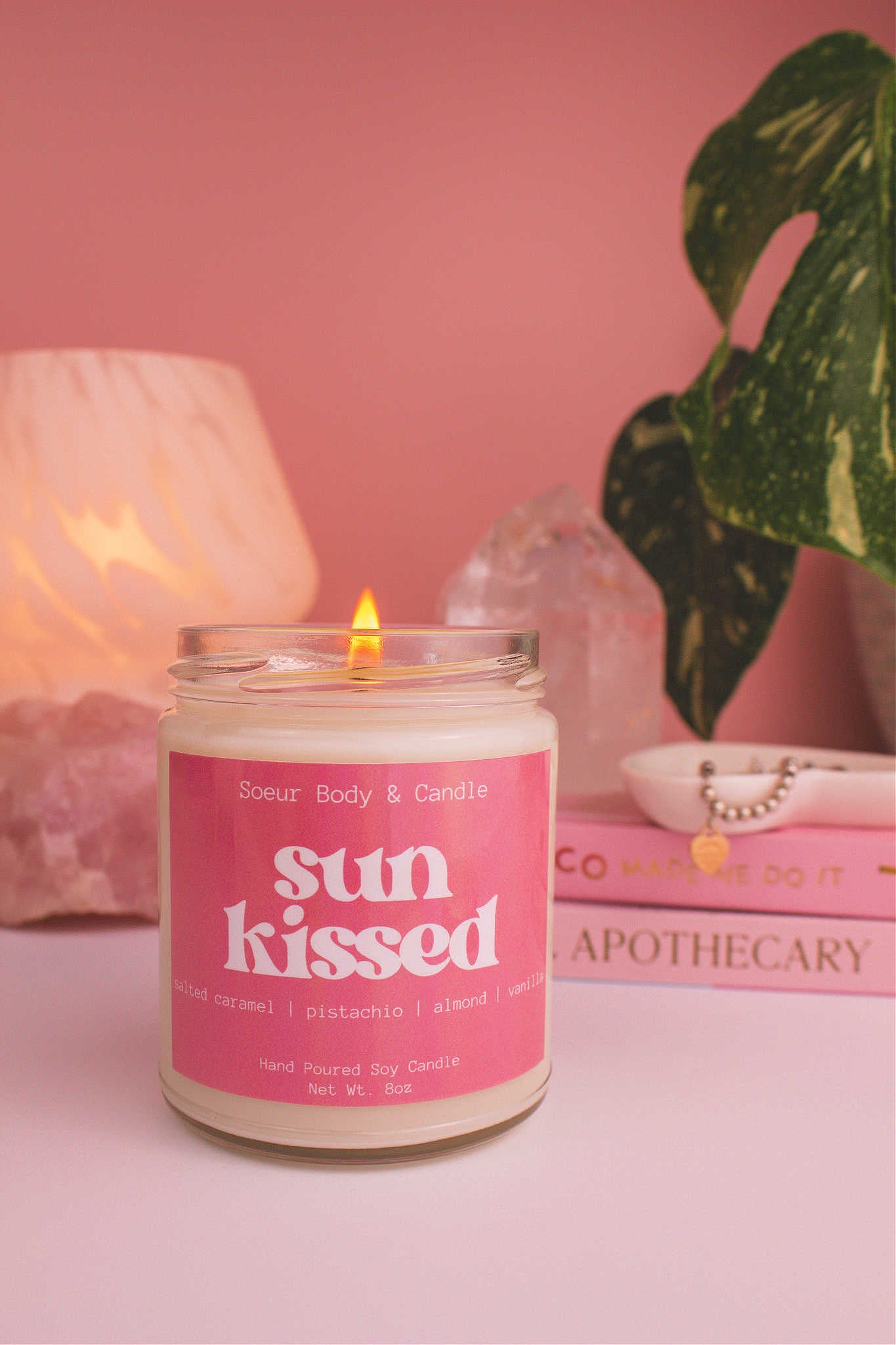 Sun Kissed candle lit, with a stack of books and a jewelry dish in the background, set against a lamp, plant, and crystals; fragrance notes: salted caramel, pistachio, almond, and vanilla.