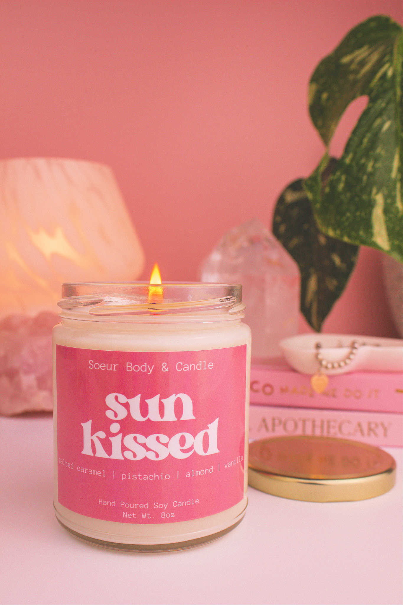 Close-up of Sun Kissed candle lit, with a stack of books and a jewelry dish in the background, set against a lamp, plant, and crystals; fragrance notes: salted caramel, pistachio, almond, and vanilla.