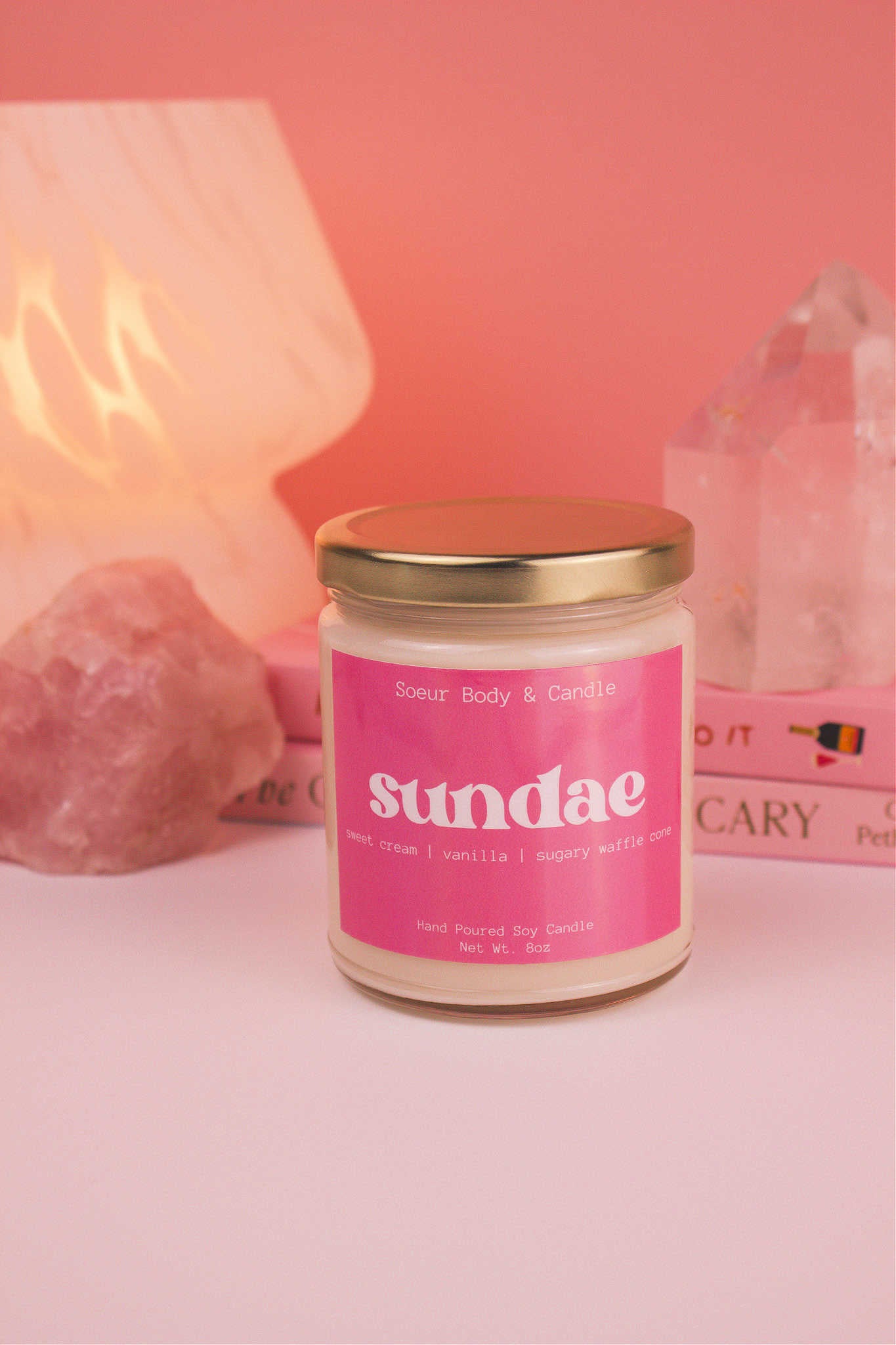 Sundae candle with the lid on, with a stack of books, a lamp, and crystals in the background; fragrance notes: sweet cream, vanilla, and sugary waffle cone.