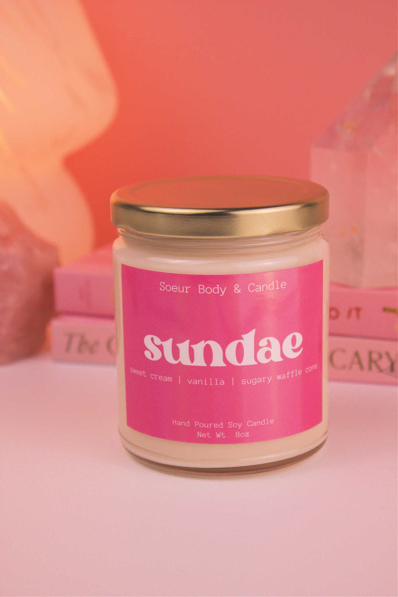 Close-up of Sundae candle with the lid on, with a stack of books, a lamp, and crystals in the background; fragrance notes: sweet cream, vanilla, and sugary waffle cone.