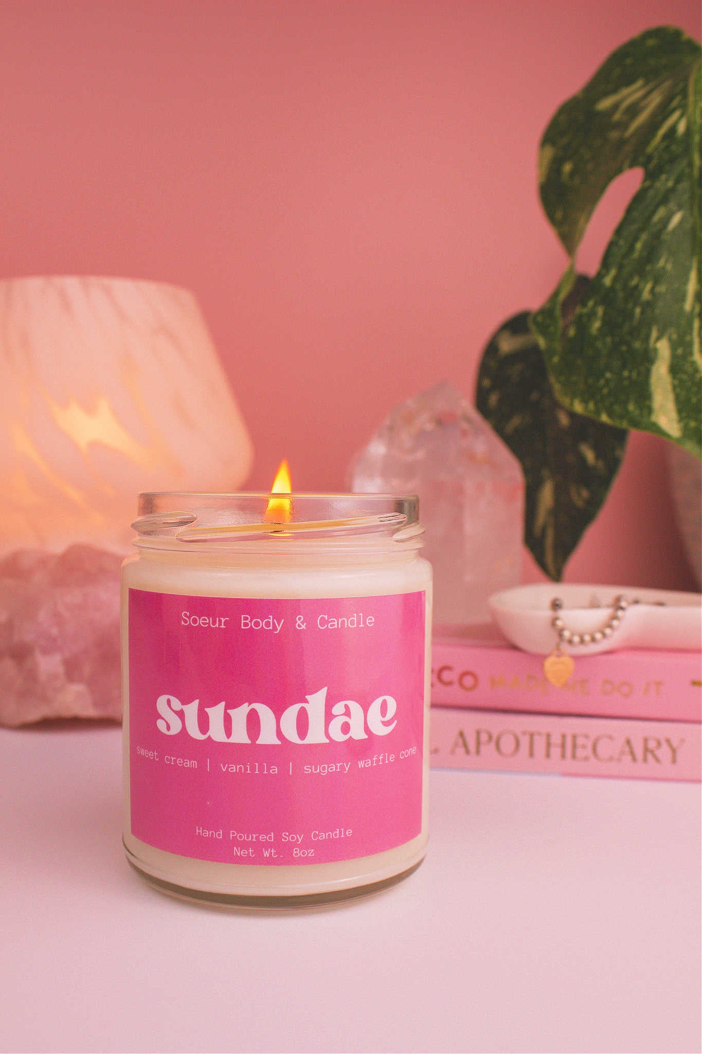 Sundae candle lit, with a stack of books and a jewelry dish in the background, set against a lamp, plant, and crystals; fragrance notes: sweet cream, vanilla, and sugary waffle cone.