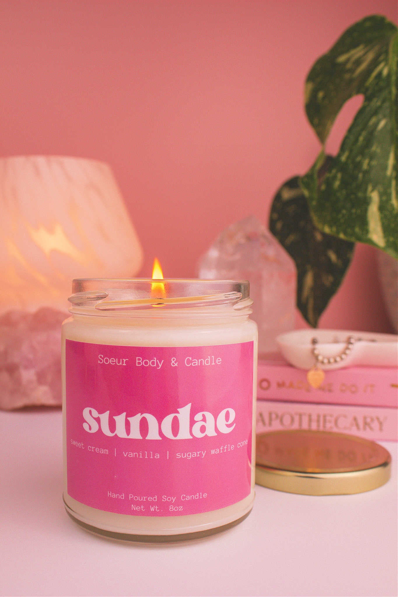 Close-up of Sundae candle lit, with a stack of books and a jewelry dish in the background, set against a lamp, plant, and crystals; fragrance notes: sweet cream, vanilla, and sugary waffle cone.