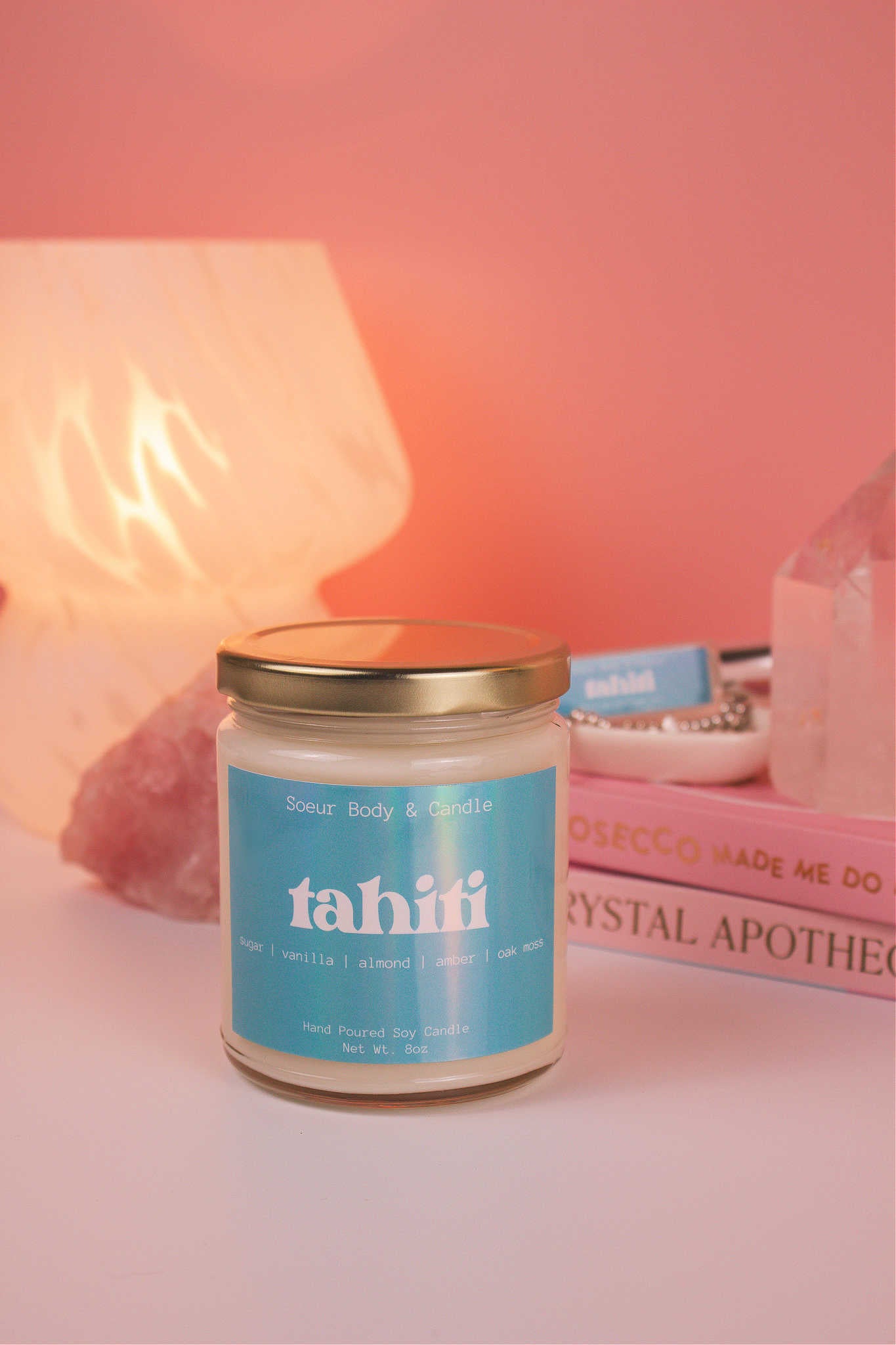 Tahiti candle with the lid on, with a stack of books and a jewelry dish in the background, set against a lamp and crystals; fragrance notes: sugar, vanilla, almond, amber, and oak moss.