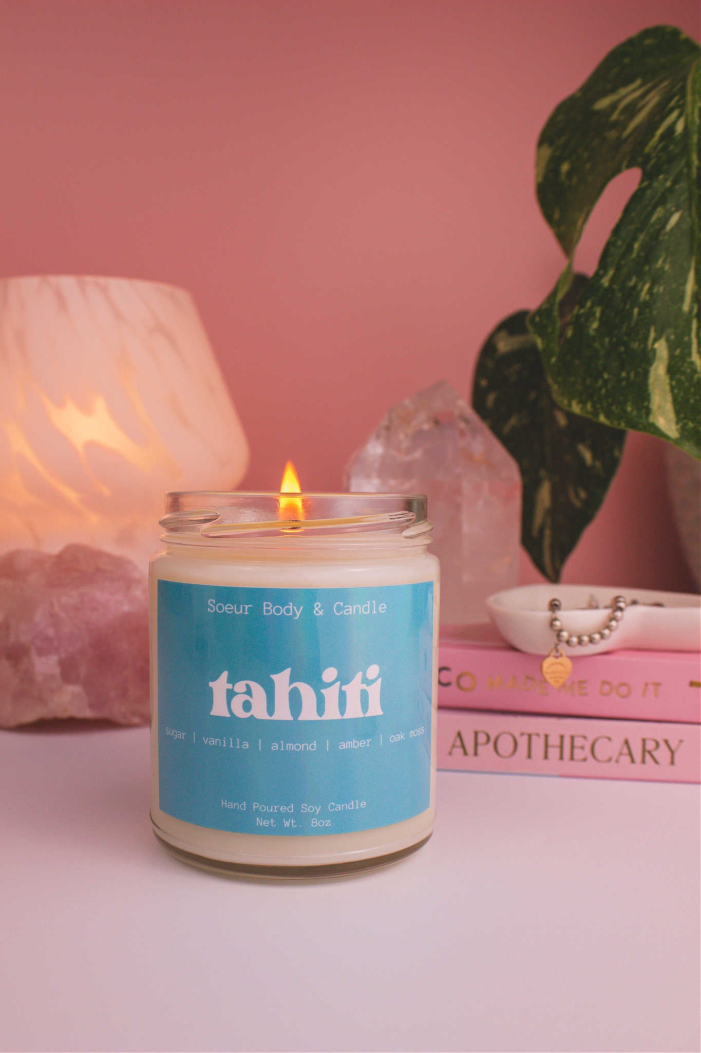Tahiti candle lit, with a stack of books and a jewelry dish in the background, set against a lamp, plant, and crystals; fragrance notes: sugar, vanilla, almond, amber, and oak moss.