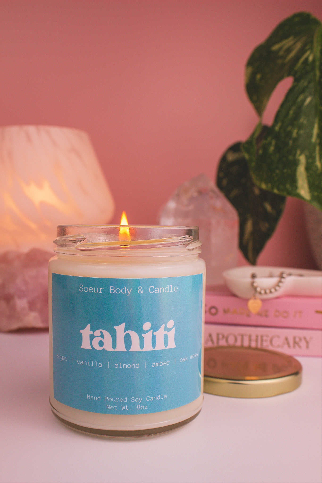Close-up of Tahiti candle lit, with a stack of books and a jewelry dish in the background, set against a lamp, plant, and crystals; fragrance notes: sugar, vanilla, almond, amber, and oak moss.