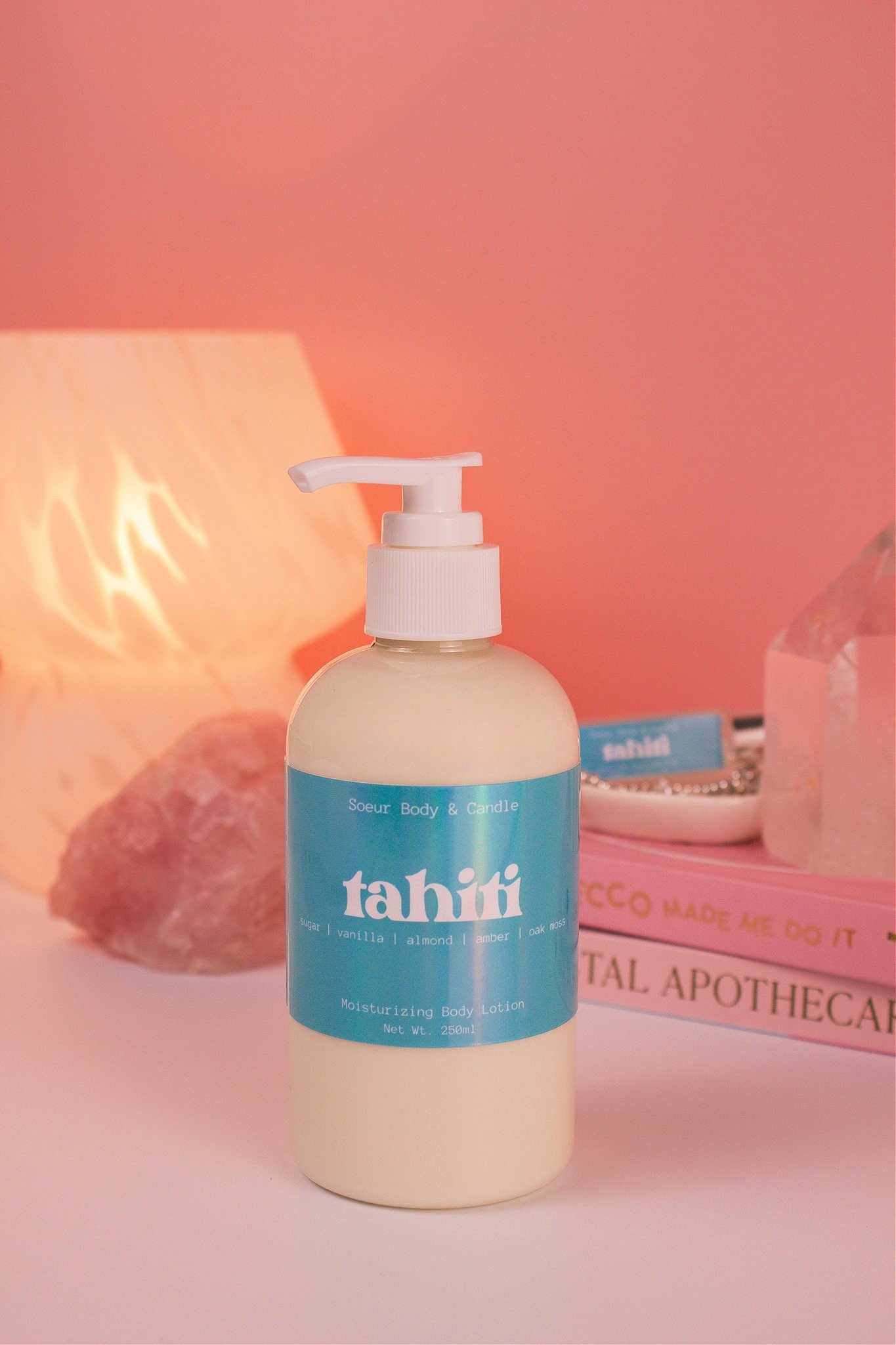 Tahiti body lotion with a stack of books, a jewelry dish, lamp, and crystals in the background; fragrance notes: sugar, vanilla, almond, amber, and oak moss.