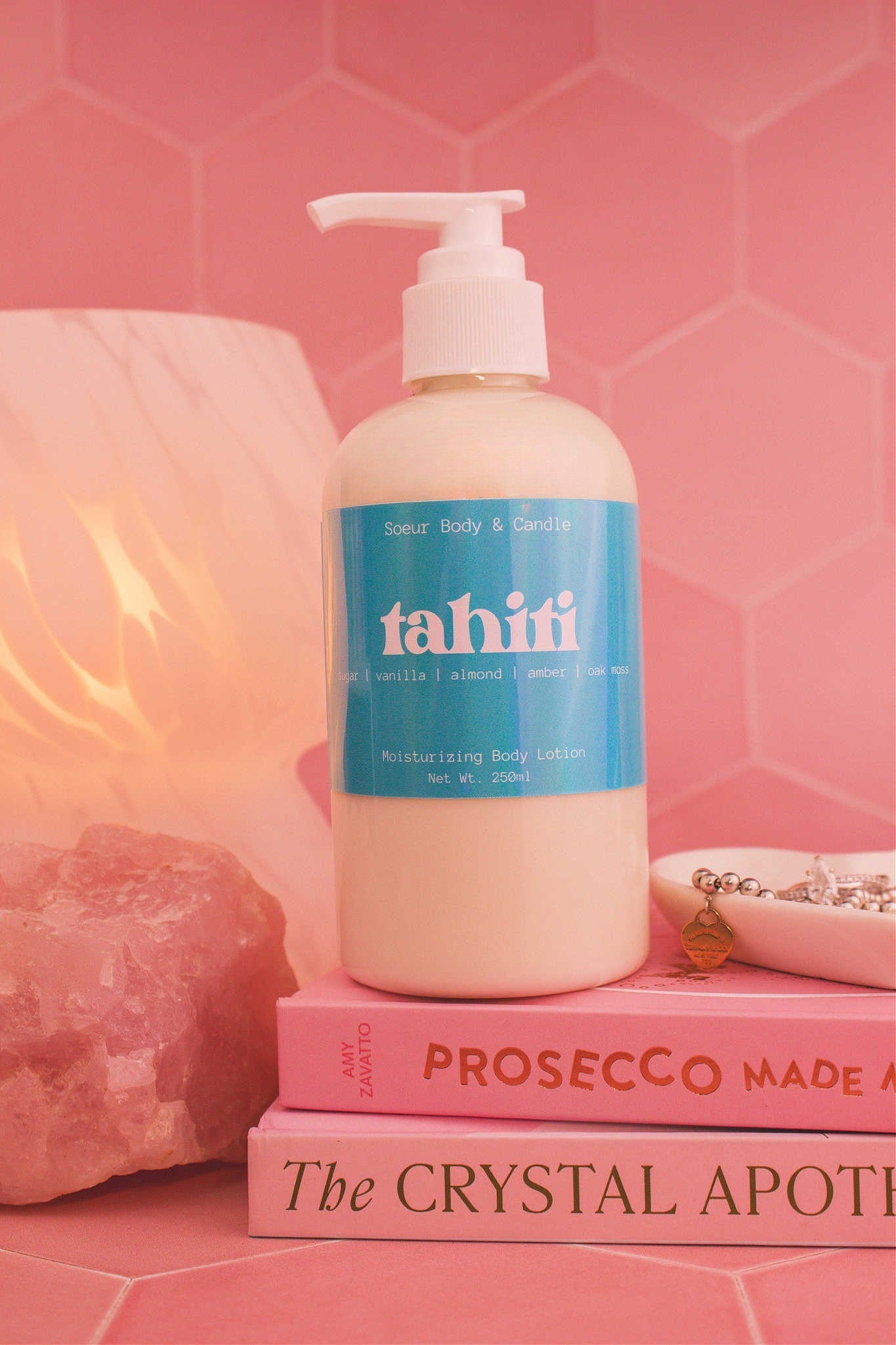 Close-up of the Tahiti body lotion on a stack of books with a jewelry dish, lamp, and crystal in the background; fragrance notes: sugar, vanilla, almond, amber, and oak moss.