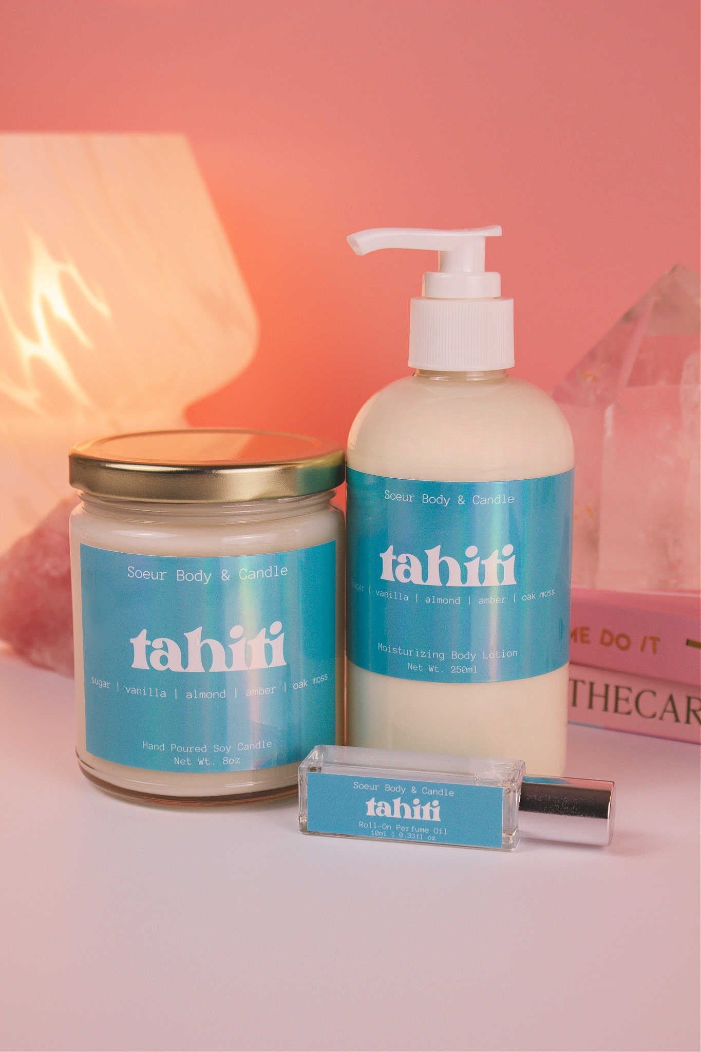 Tahiti body lotion presented with a matching candle and roll-on perfume, with books, a lamp, and crystals in the background; highlighting sugar, vanilla, almond, amber, and oak moss notes.